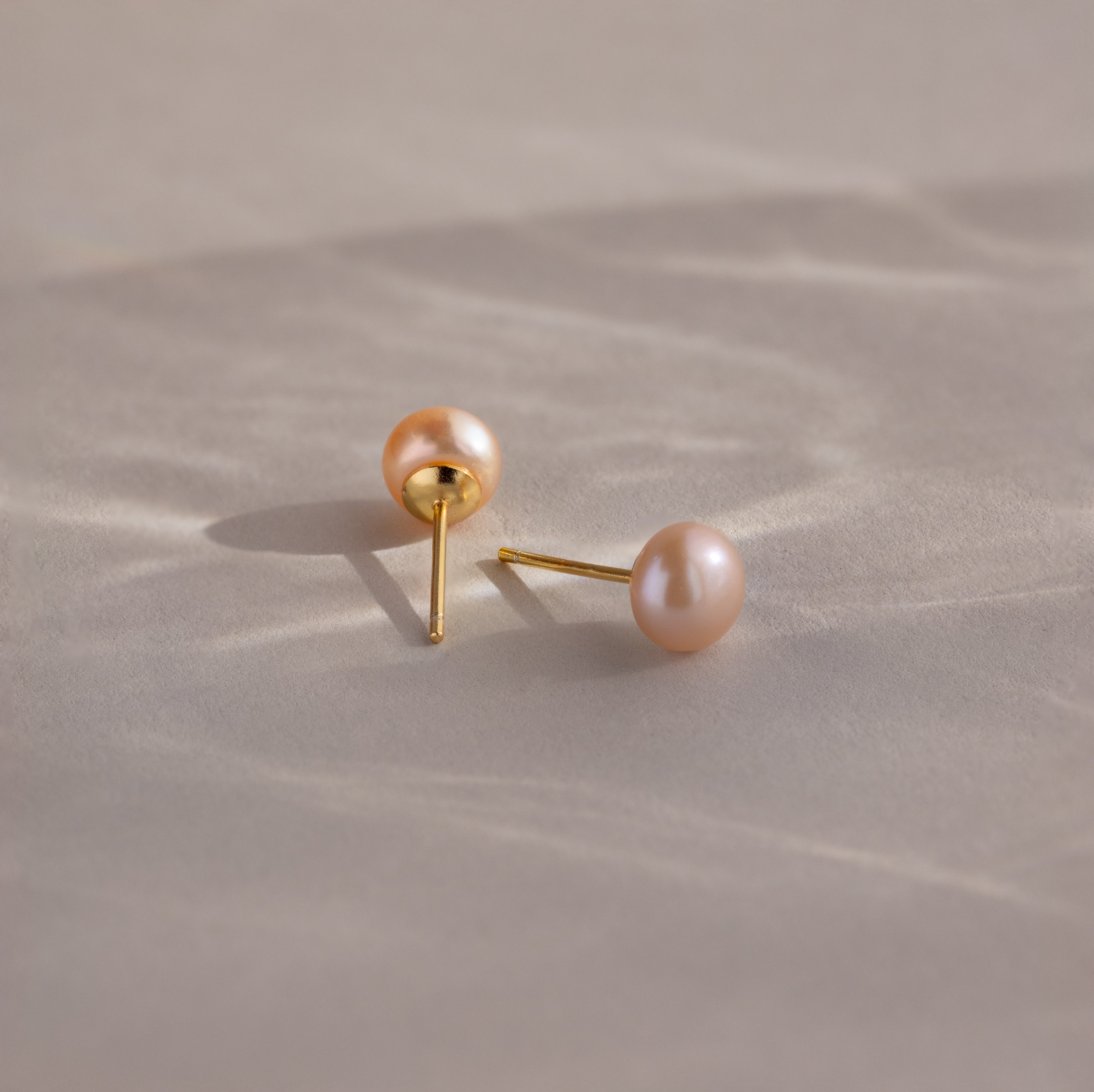 A pair of Pink Pearl Studs in 18K Gold is displayed on a soft, blush-toned surface with gentle shadows.