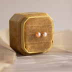A pair of Pink Pearl Studs in Sterling Silver displayed on a textured beige jewelry box.