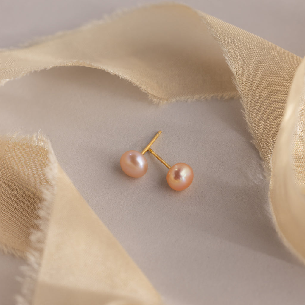 Pink Pearl Studs featuring blush tone freshwater pearls are displayed on a beige surface, accented by sheer beige fabric strips.