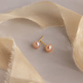Pink Pearl Studs featuring blush tone freshwater pearls are displayed on a beige surface, accented by sheer beige fabric strips.