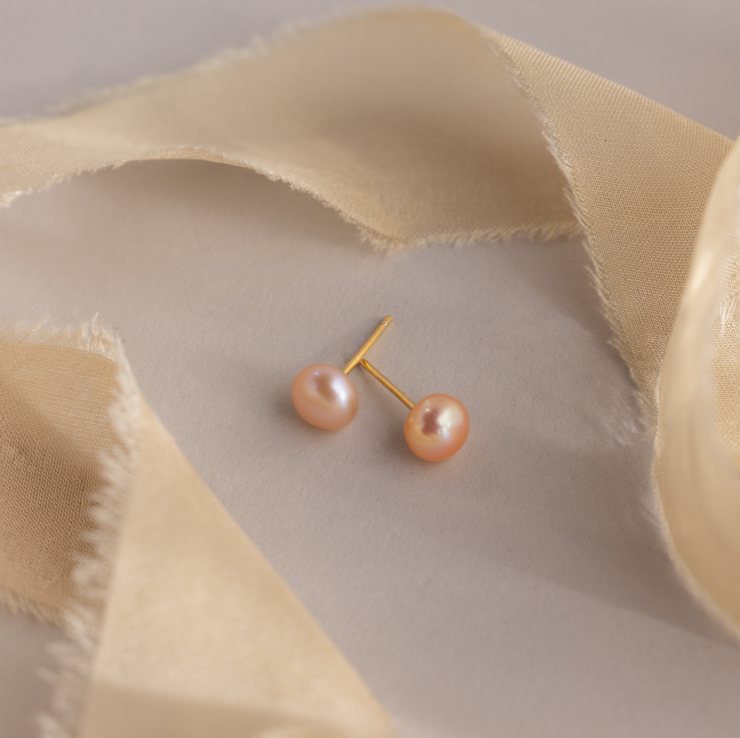 Pink Pearl Studs featuring blush tone freshwater pearls are displayed on a beige surface, accented by sheer beige fabric strips.