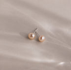 Pink Pearl Studs in a blush tone are showcased elegantly against a soft, light beige satin fabric background.