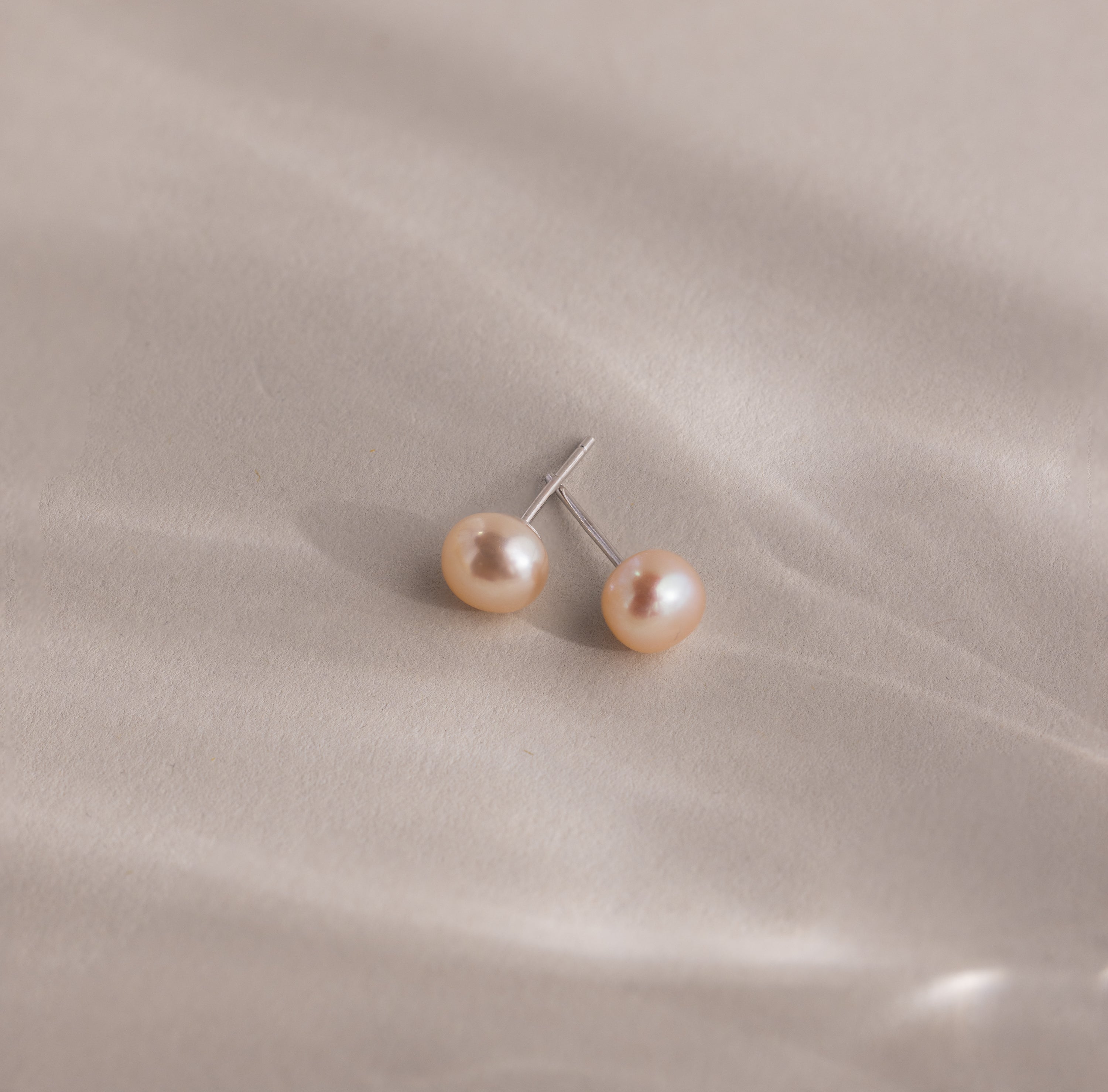 Pink Pearl Studs in a blush tone are showcased elegantly against a soft, light beige satin fabric background.