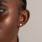 A close-up shows a person's ear wearing a Pink Pearl Stud in Sterling Silver, featuring a blush-tone freshwater pearl, with part of their face visible.