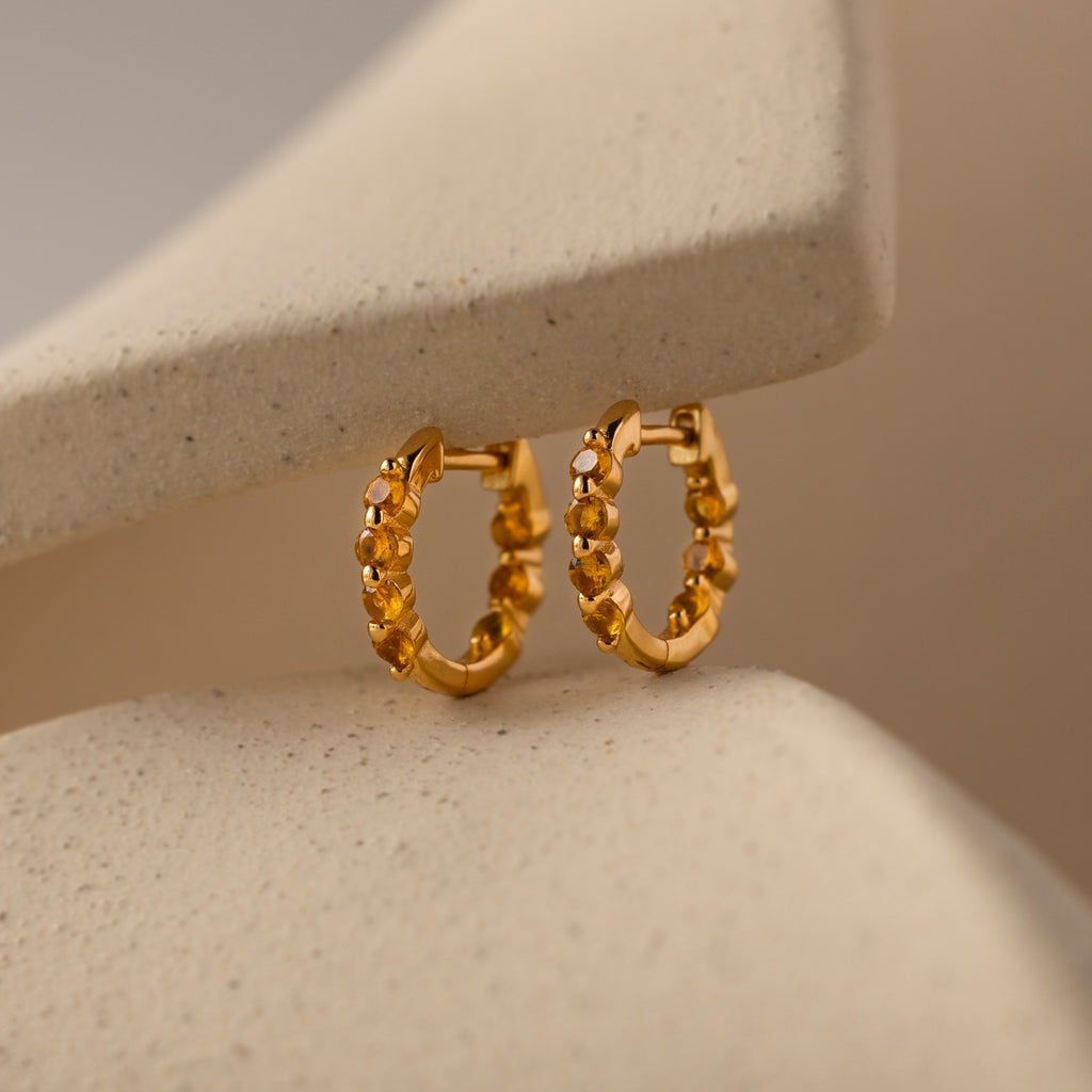 Mini Birthstone Beaded Huggies with a textured design shine against a soft beige background—add a unique touch of birthstone jewelry elegance to your look.