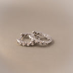 Two Mini Birthstone Beaded Huggies—silver hoops with small embedded stones—are displayed on a soft beige surface.