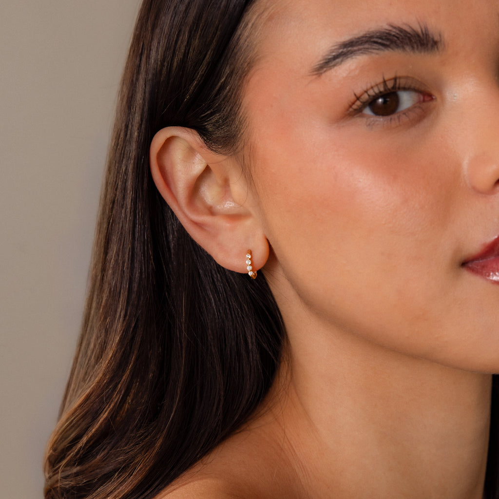 Woman with long dark hair wears Mini Topaz Beaded Huggies—modern small gold hoops with beaded details—shown from the side.