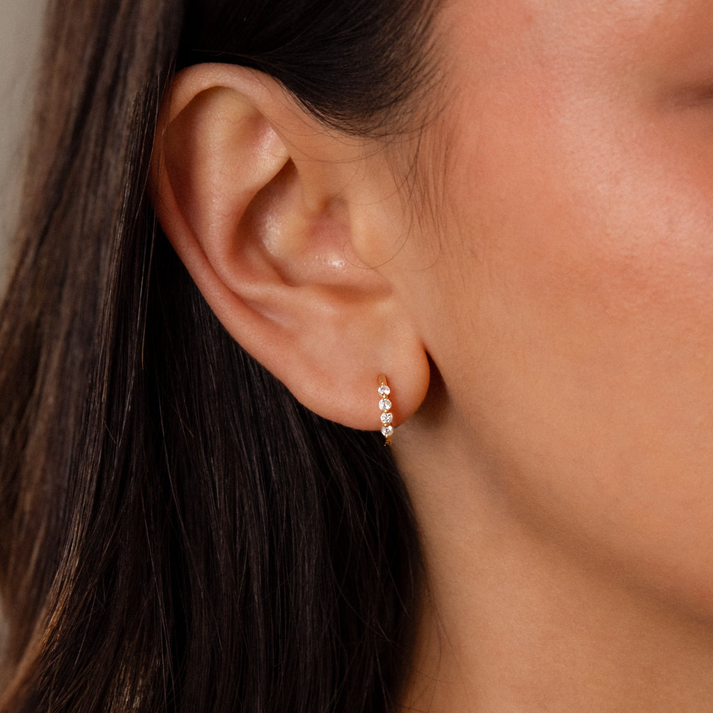 Close-up of a womans ear wearing Mini Birthstone Beaded Huggies—dainty gold earrings accented with three sparkling birthstones for a subtle, elegant touch.