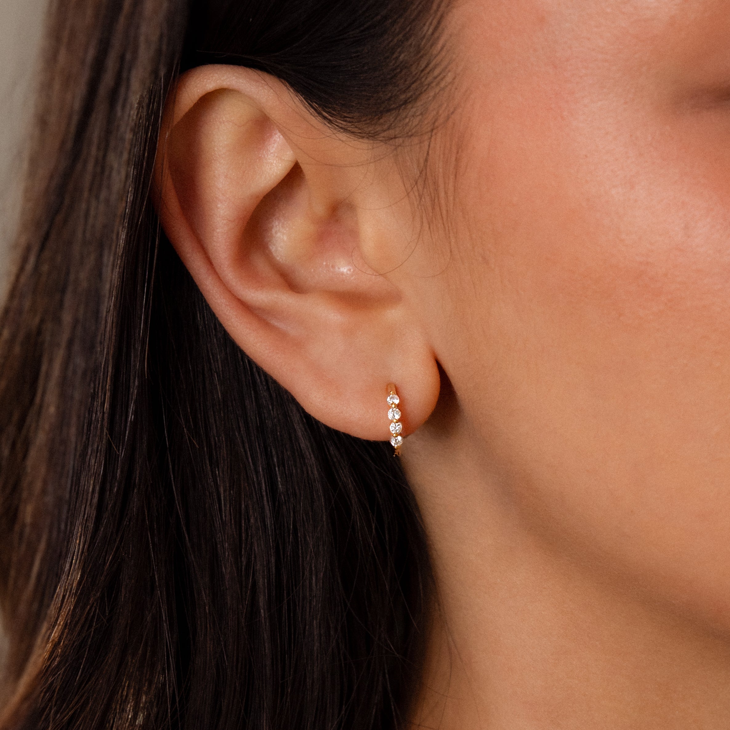 Close-up of a womans ear wearing Mini Birthstone Beaded Huggies—dainty gold earrings accented with three sparkling birthstones for a subtle, elegant touch.