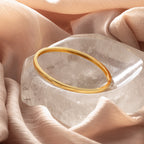 The Classic Bangle in 18K Gold rests on a clear crystal and soft peach fabric—a chic, minimalist accessory perfect for those who love elegant jewelry.