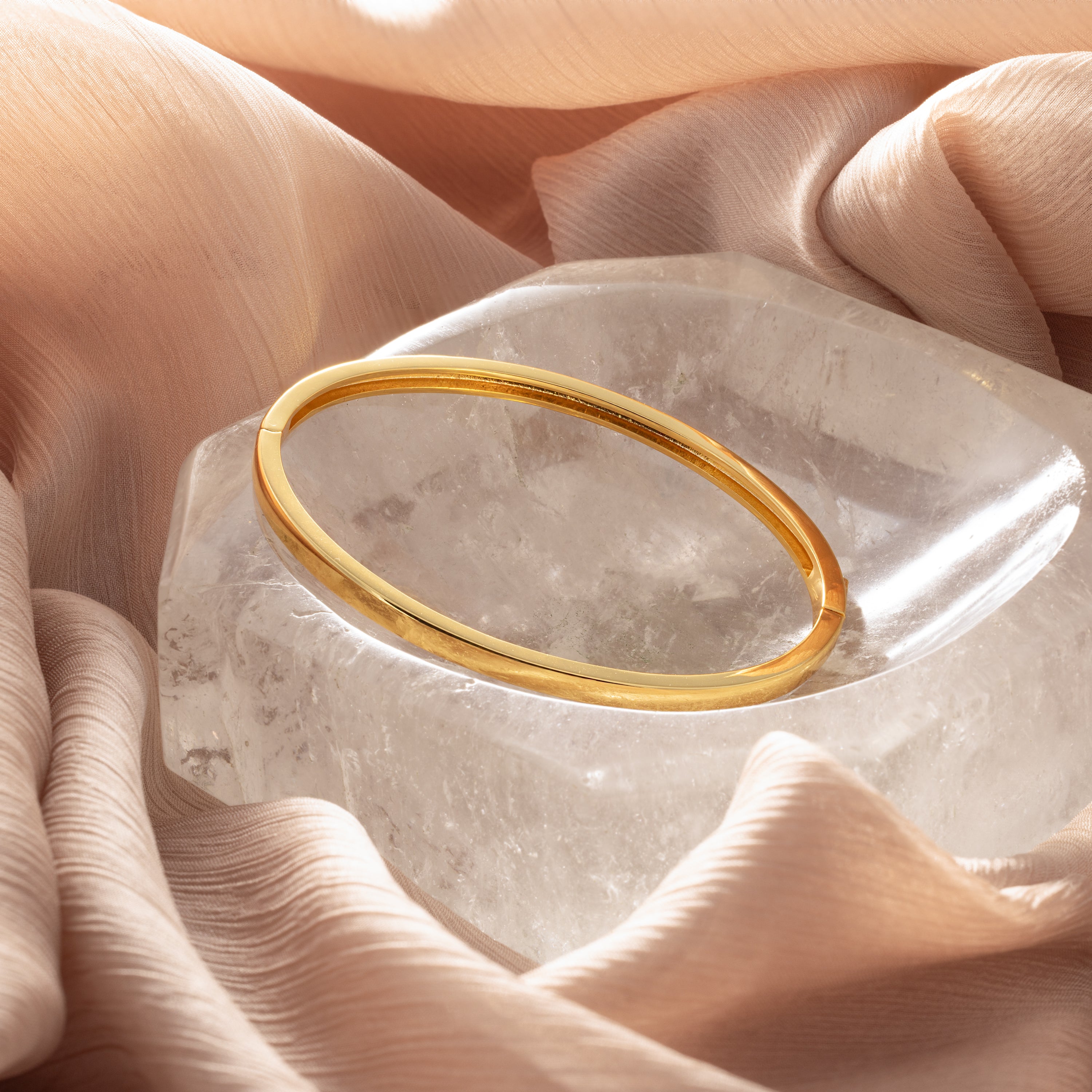 The Classic Bangle in 18K Gold rests on a clear crystal and soft peach fabric—a chic, minimalist accessory perfect for those who love elegant jewelry.
