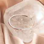 The Classic Bangle in Sterling Silver adds a chic touch, displayed on a large clear crystal and nestled among soft beige fabric folds.