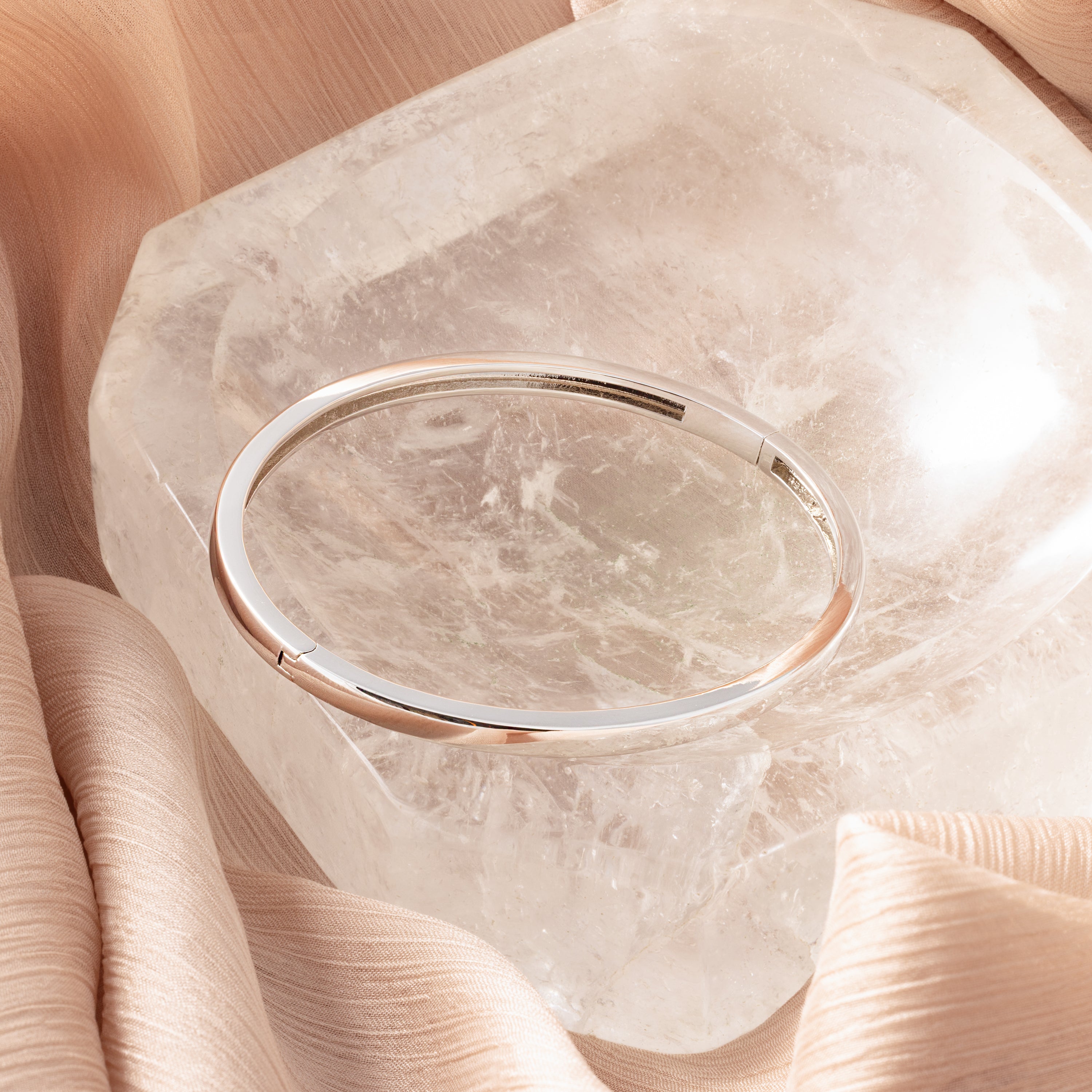 The Classic Bangle in Sterling Silver adds a chic touch, displayed on a large clear crystal and nestled among soft beige fabric folds.