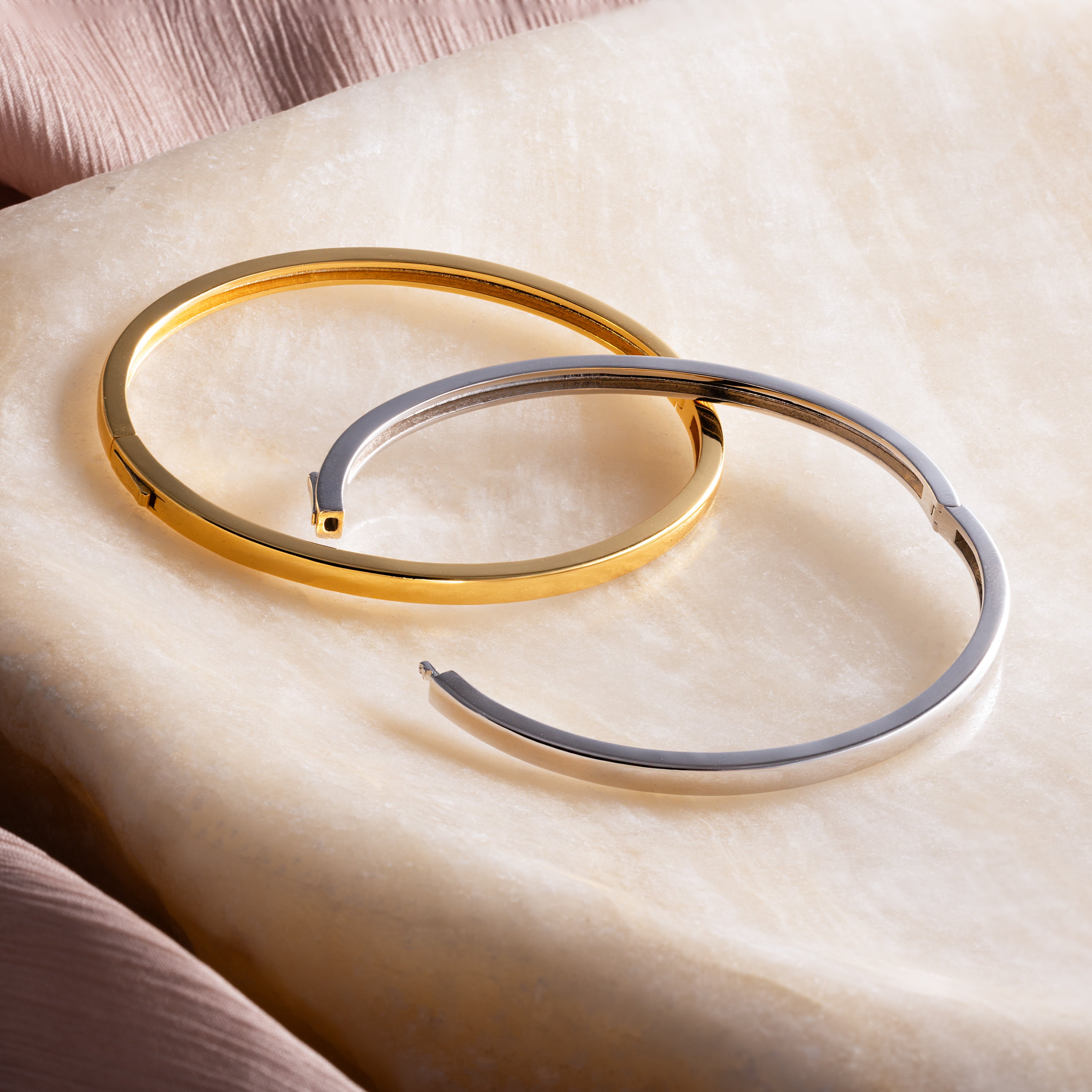 The Classic Bangle in 18K Gold is displayed on marble with soft pink fabric—a minimalist, chic jewelry piece that elevates any look.