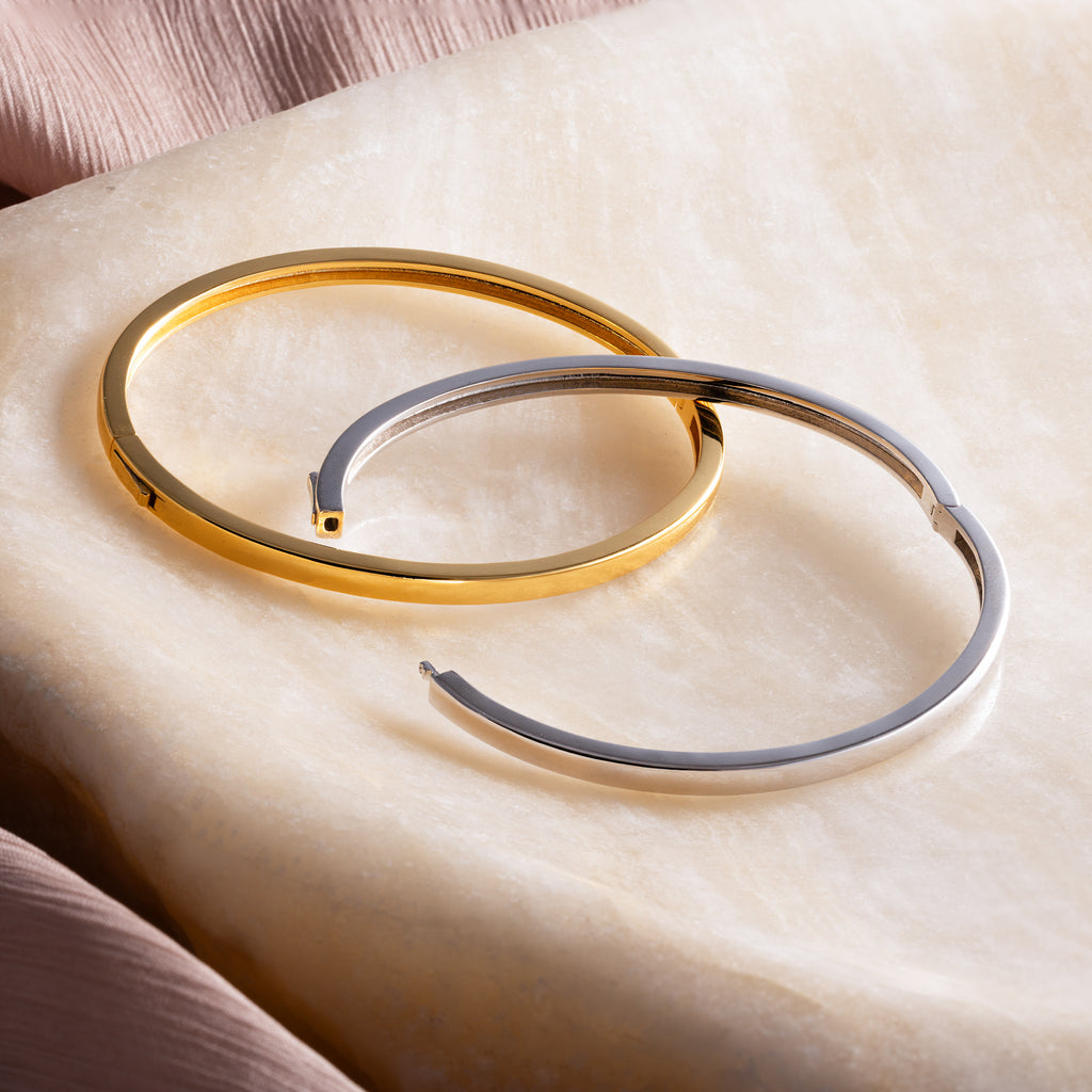 The Classic Bangle, available in gold and silver, rests on smooth marble beside soft pink fabric, bringing chic elegance to your jewelry collection.