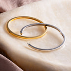 The Classic Bangle, available in gold and silver, rests on smooth marble beside soft pink fabric, bringing chic elegance to your jewelry collection.