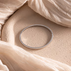 The Classic Bangle in Sterling Silver, a chic minimalist jewelry piece, is displayed on a textured beige surface with sheer, flowing fabric draped around it.