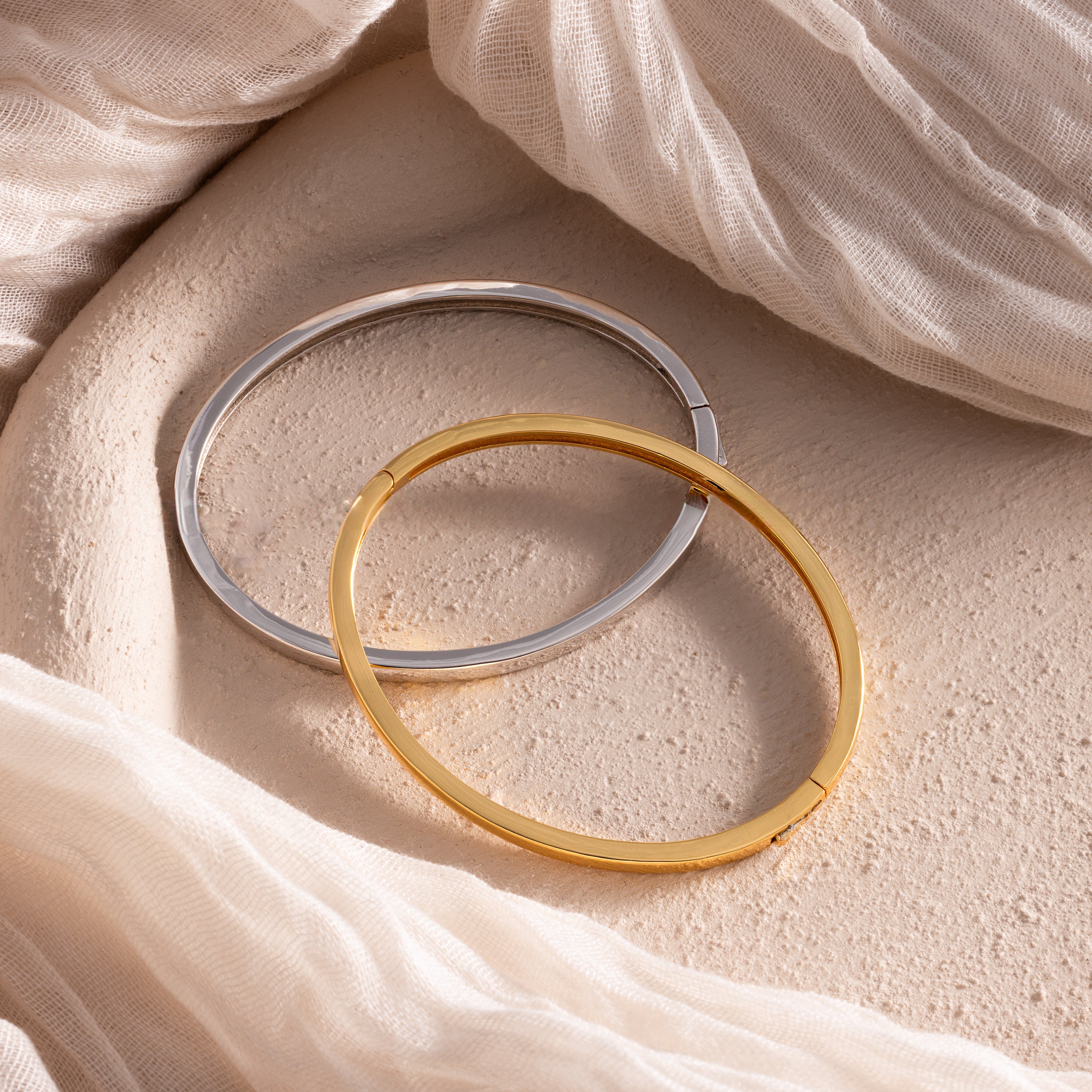 The Classic Bangle in 18K Gold rests on a textured surface with cream fabric draped nearby—a chic, minimalist accessory ideal for those who value timeless jewelry.