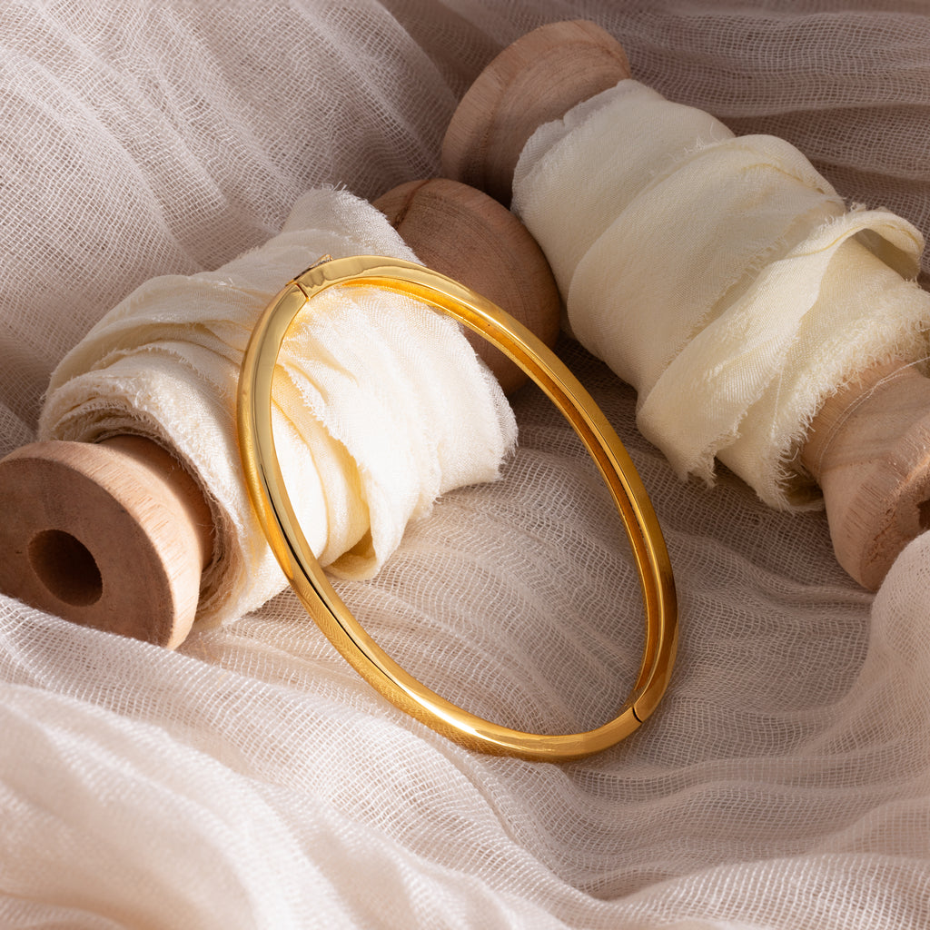 The Classic Bangle in 18K Gold rests on cream fabric-wrapped wooden spools atop a soft beige cloth, highlighting the essence of minimalist jewelry.