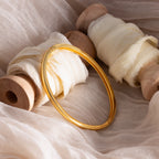 The Classic Bangle in 18K Gold rests on cream fabric-wrapped wooden spools atop a soft beige cloth, highlighting the essence of minimalist jewelry.