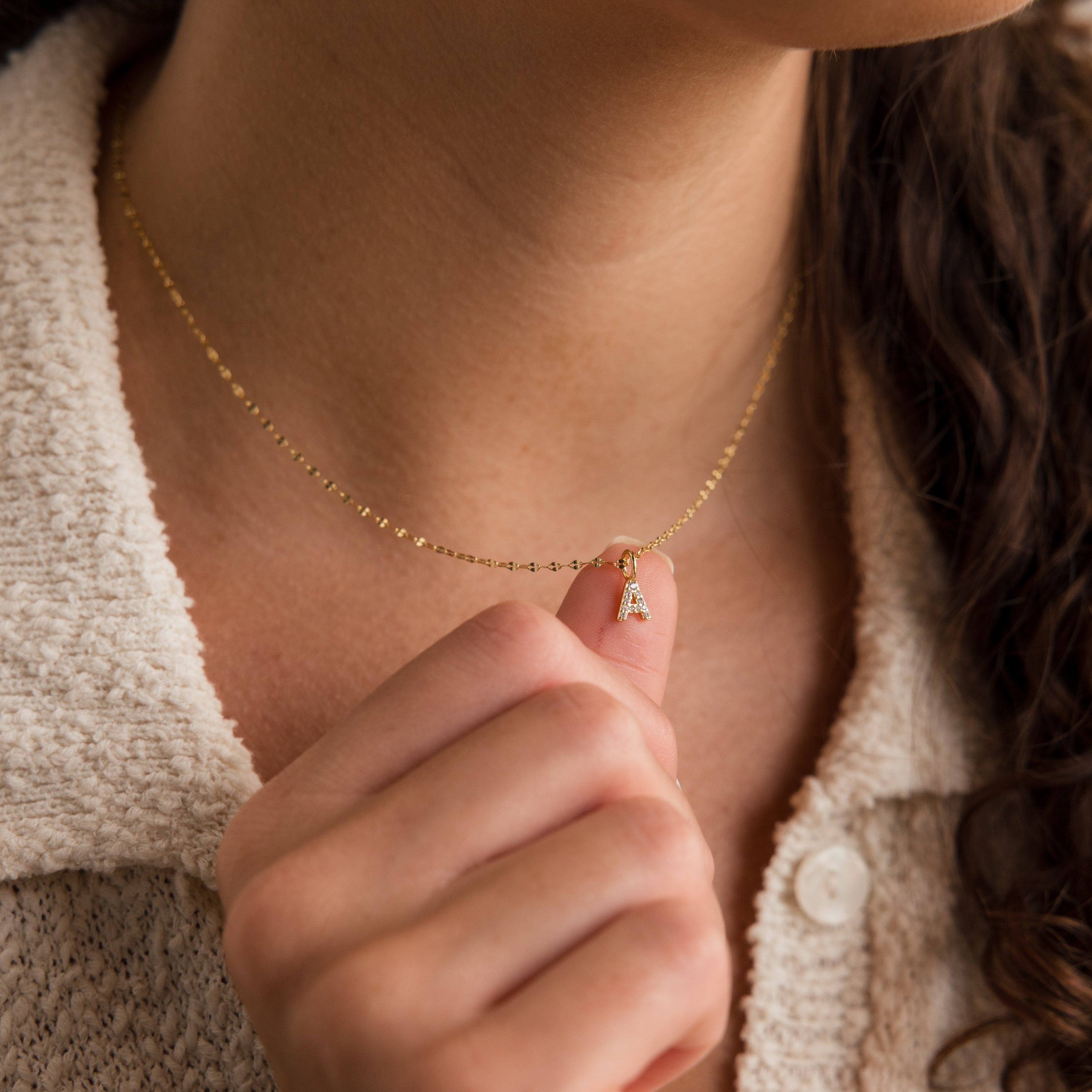 A woman wears a textured cream-colored top and holds the Dainty Pave Letter Necklace, featuring a small pavé pendant.
