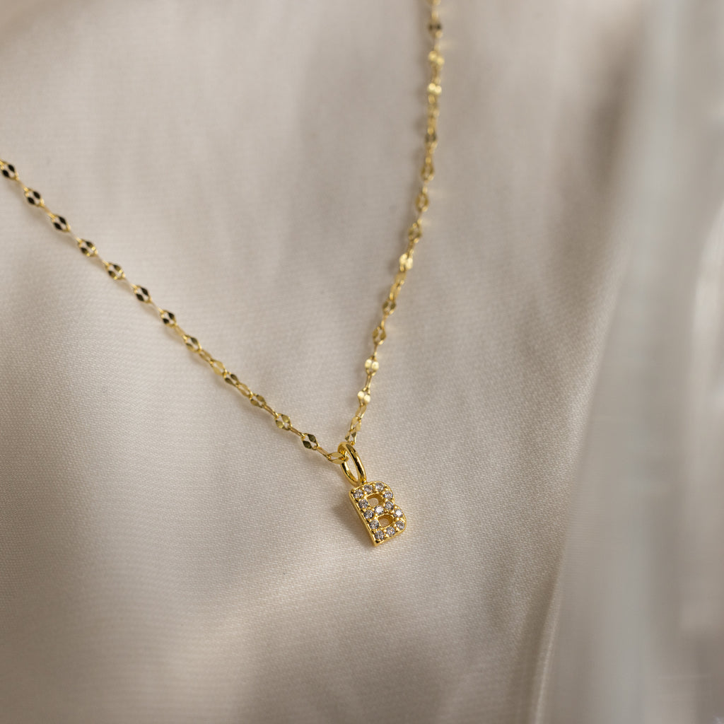The Dainty Pave Letter Necklace in 18K Gold features a pavé letter B pendant with sparkling CZ diamonds, suspended on a gold mirror chain and elegantly displayed on soft, light-colored fabric.