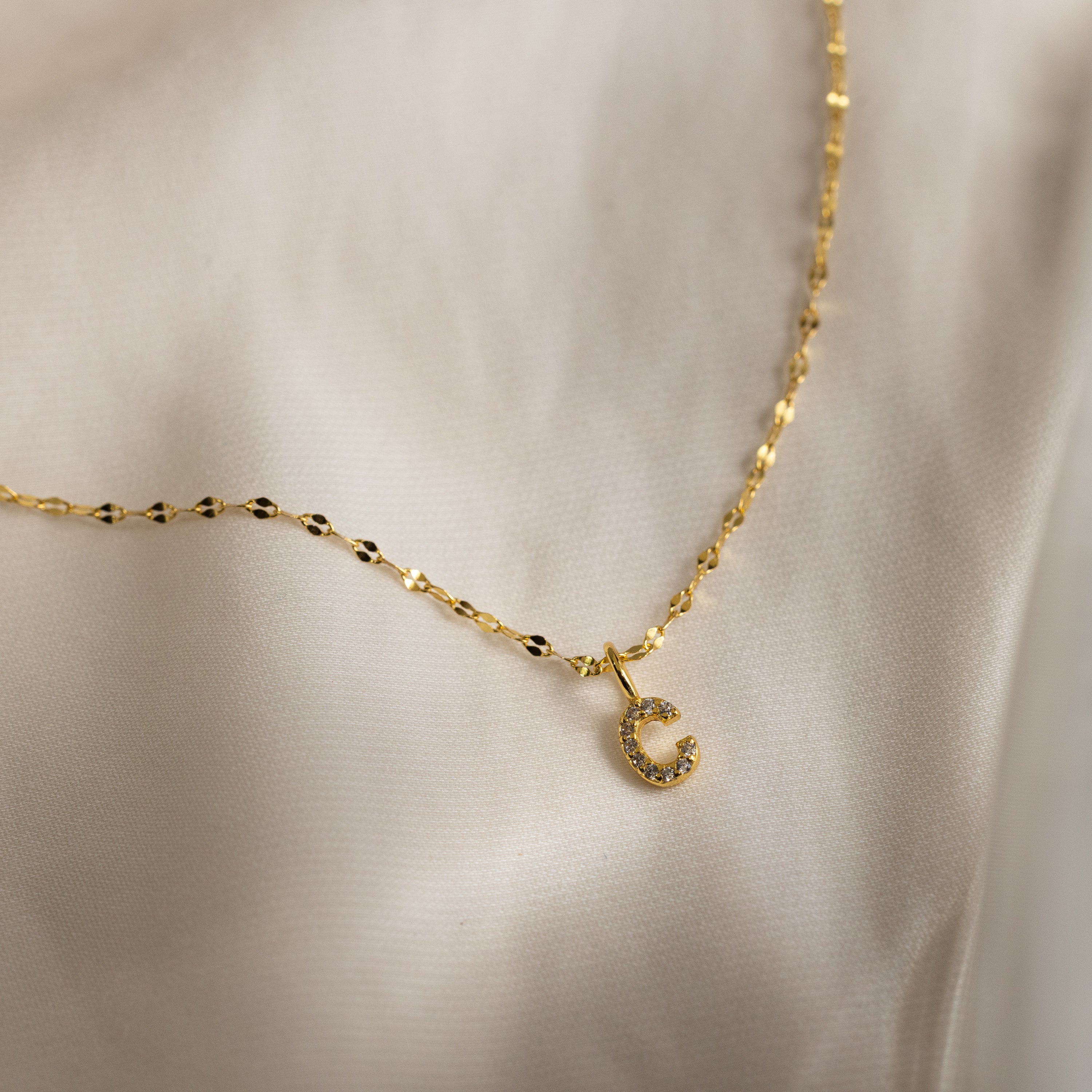 Dainty Pave Letter Necklace in 18K Gold with a pavé letter C pendant studded with sparkling CZ diamonds, shown on a cream fabric background.