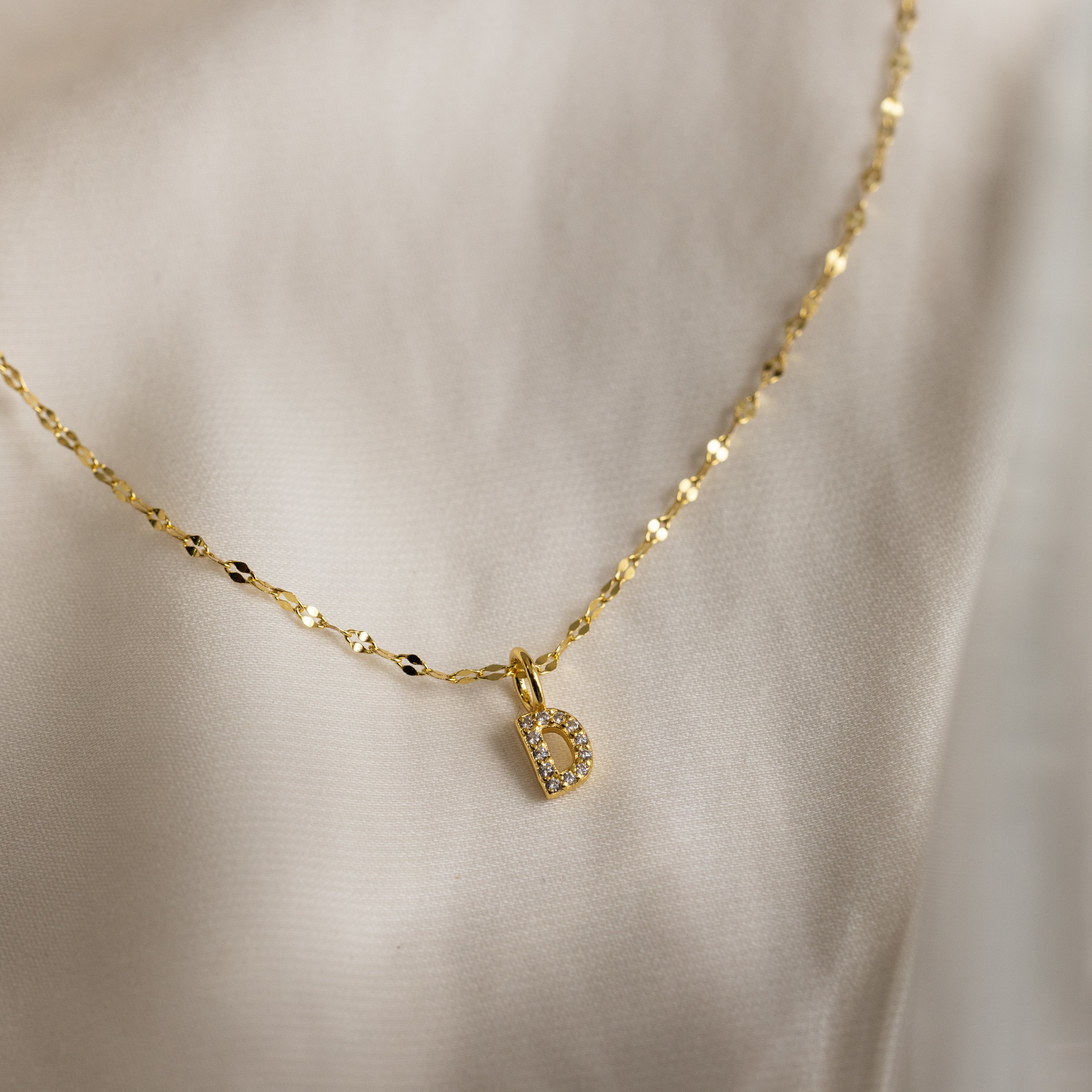 The Dainty Pave Letter Necklace in 18K Gold features a small, CZ-studded letter D pendant elegantly displayed on cream satin fabric.
