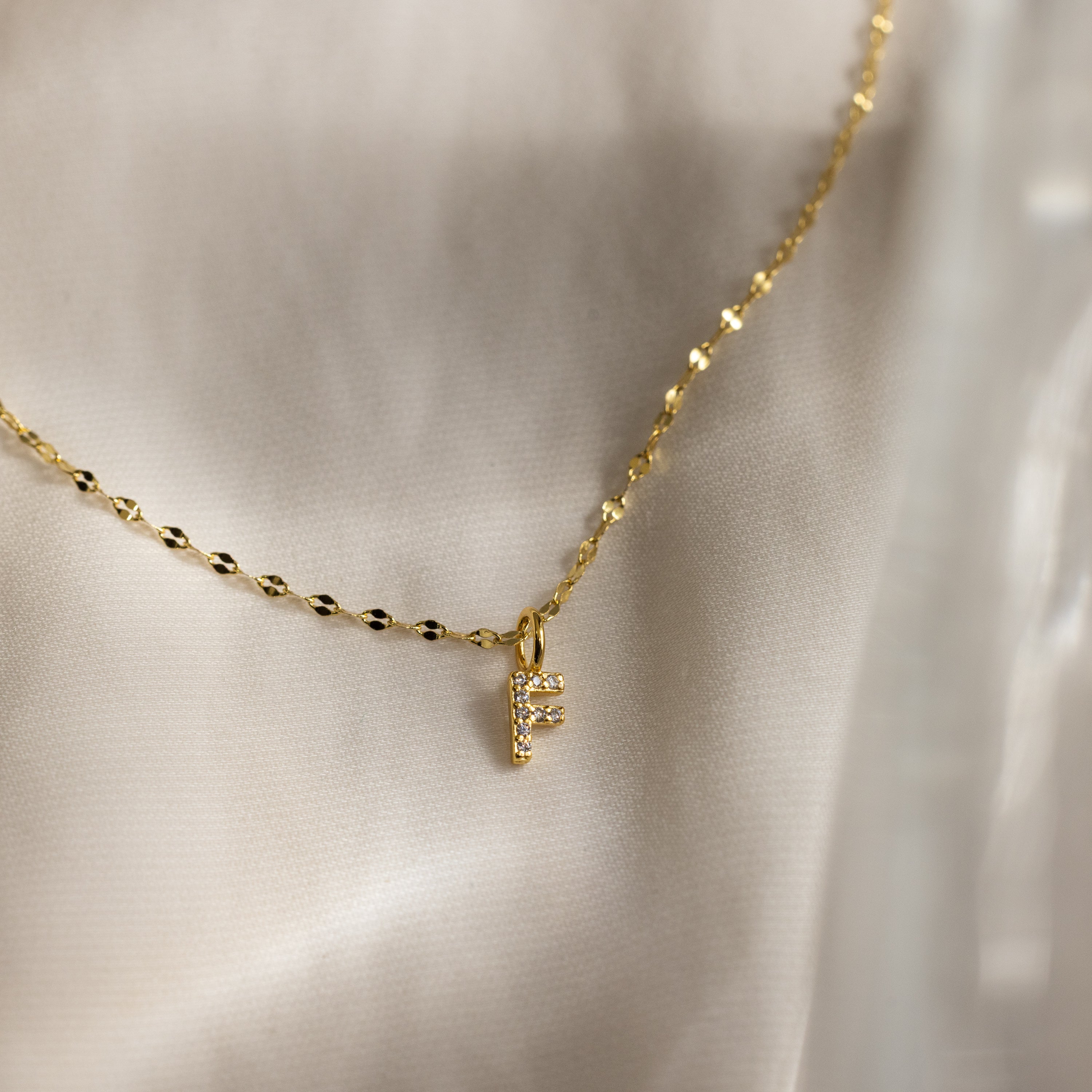 The Dainty Pave Letter Necklace in 18K Gold features a delicate gold chain and a pavé letter F pendant with sparkling CZ accents, shown on a light fabric background.
