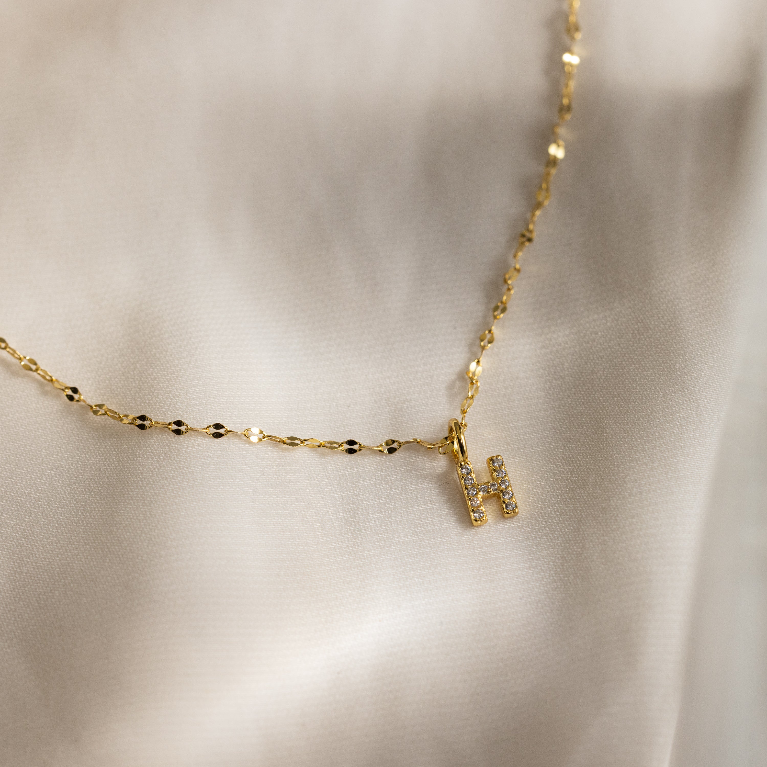 The Dainty Pave Letter Necklace in 18K Gold features a sparkly CZ-studded H pendant, beautifully displayed on a soft, silky fabric background.