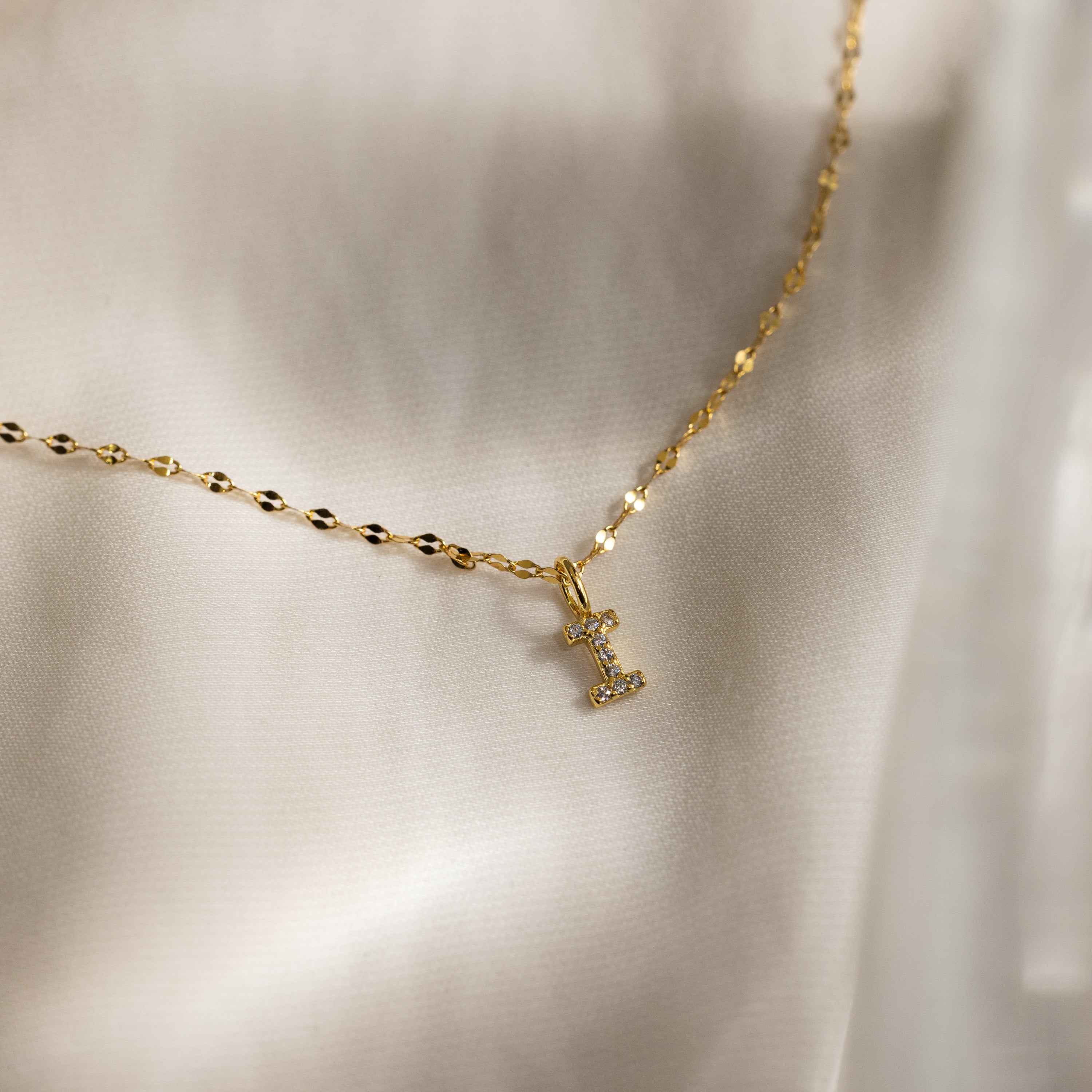 The Dainty Pave Letter Necklace in 18K Gold features a pavé letter L pendant with sparkling CZ diamonds, showcased on a light satin fabric background.