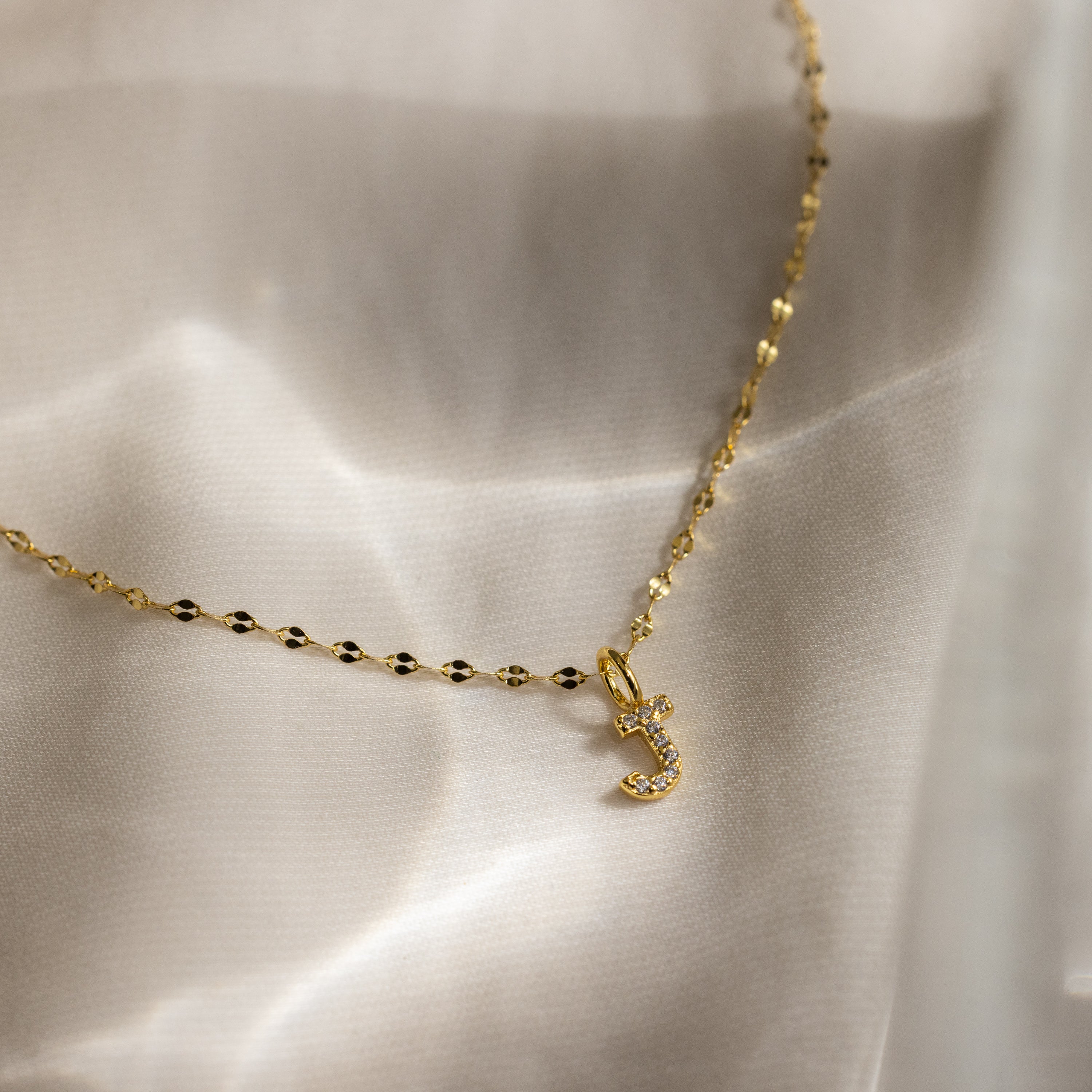 The Dainty Pave Letter Necklace in 18K Gold features a jeweled J pendant shimmering with CZ diamonds, elegantly displayed on soft ivory satin.
