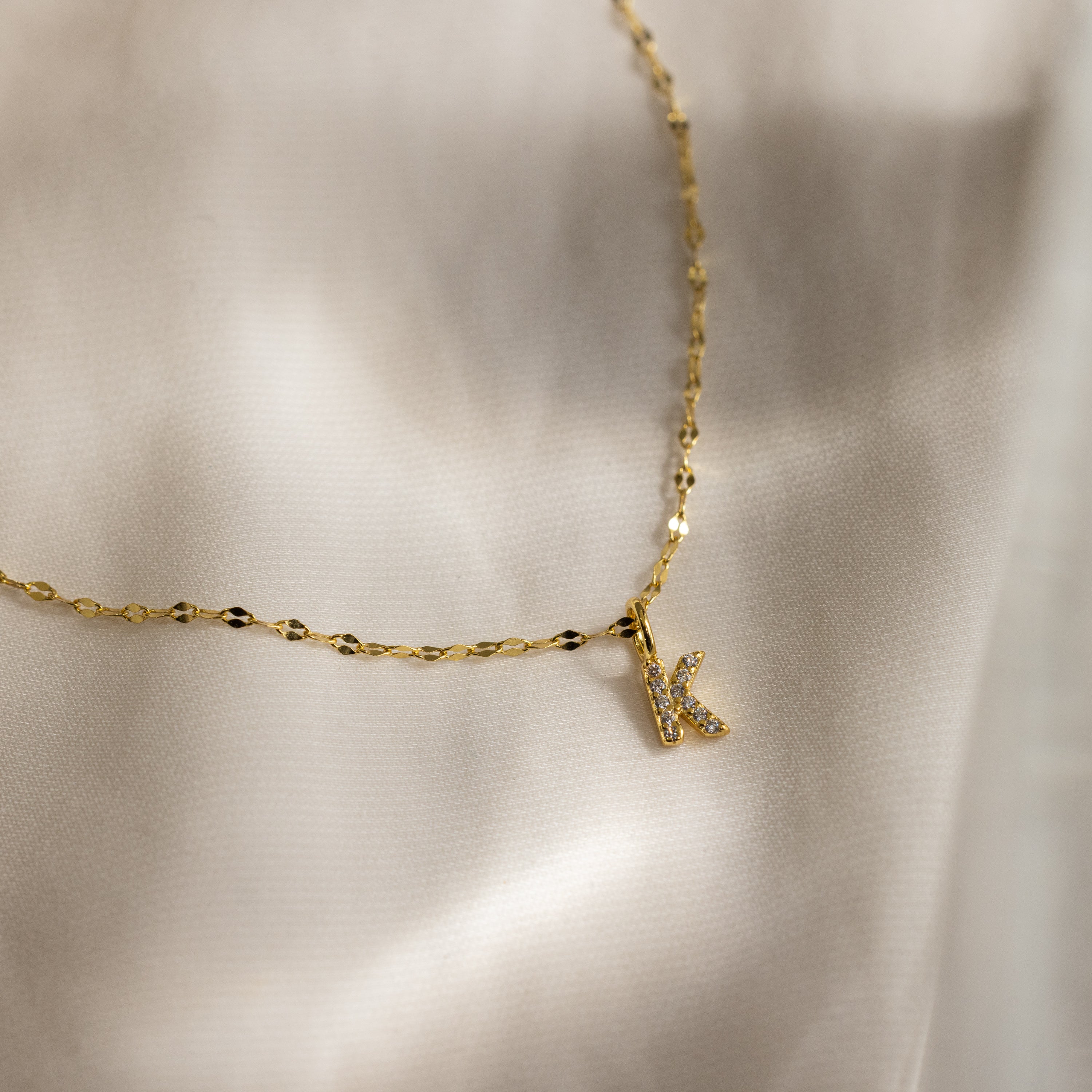 The Dainty Pave Letter Necklace in 18K Gold features a small, CZ diamond-studded K pendant resting on a soft, light-colored fabric background.