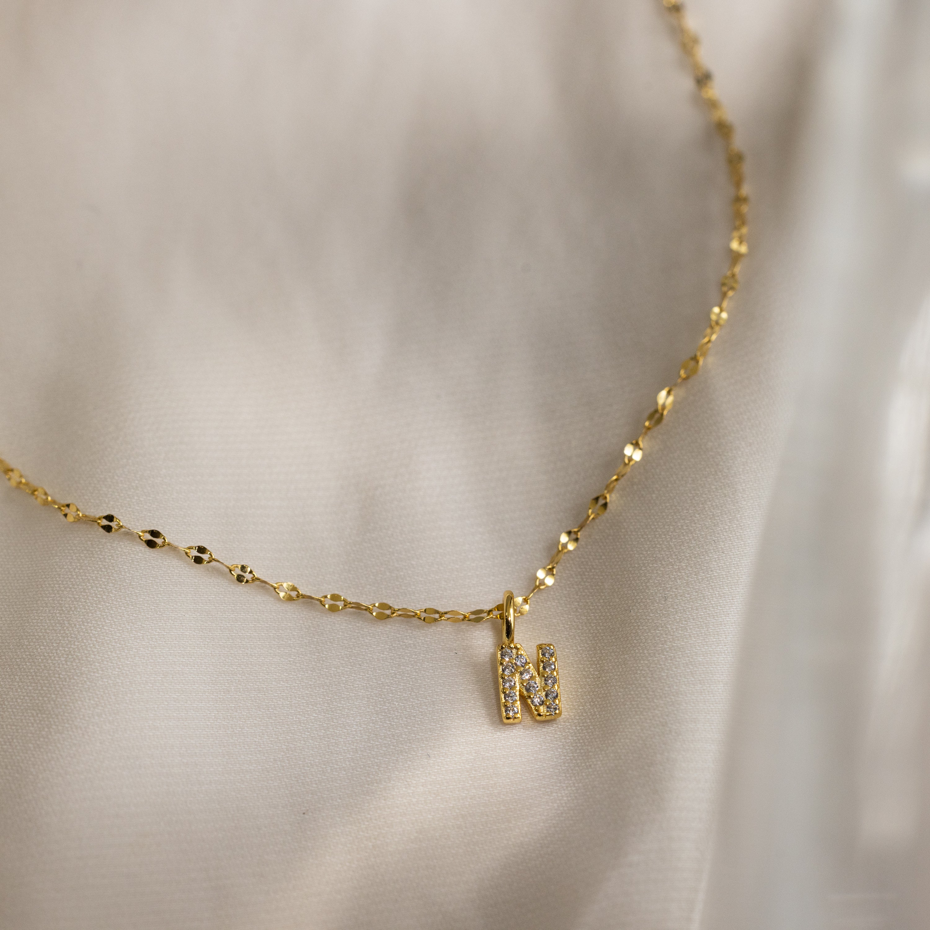 The Dainty Pave Letter Necklace in 18K Gold features a textured mirror chain and a small jeweled letter N pendant with CZ diamonds, shown here on a light fabric background.