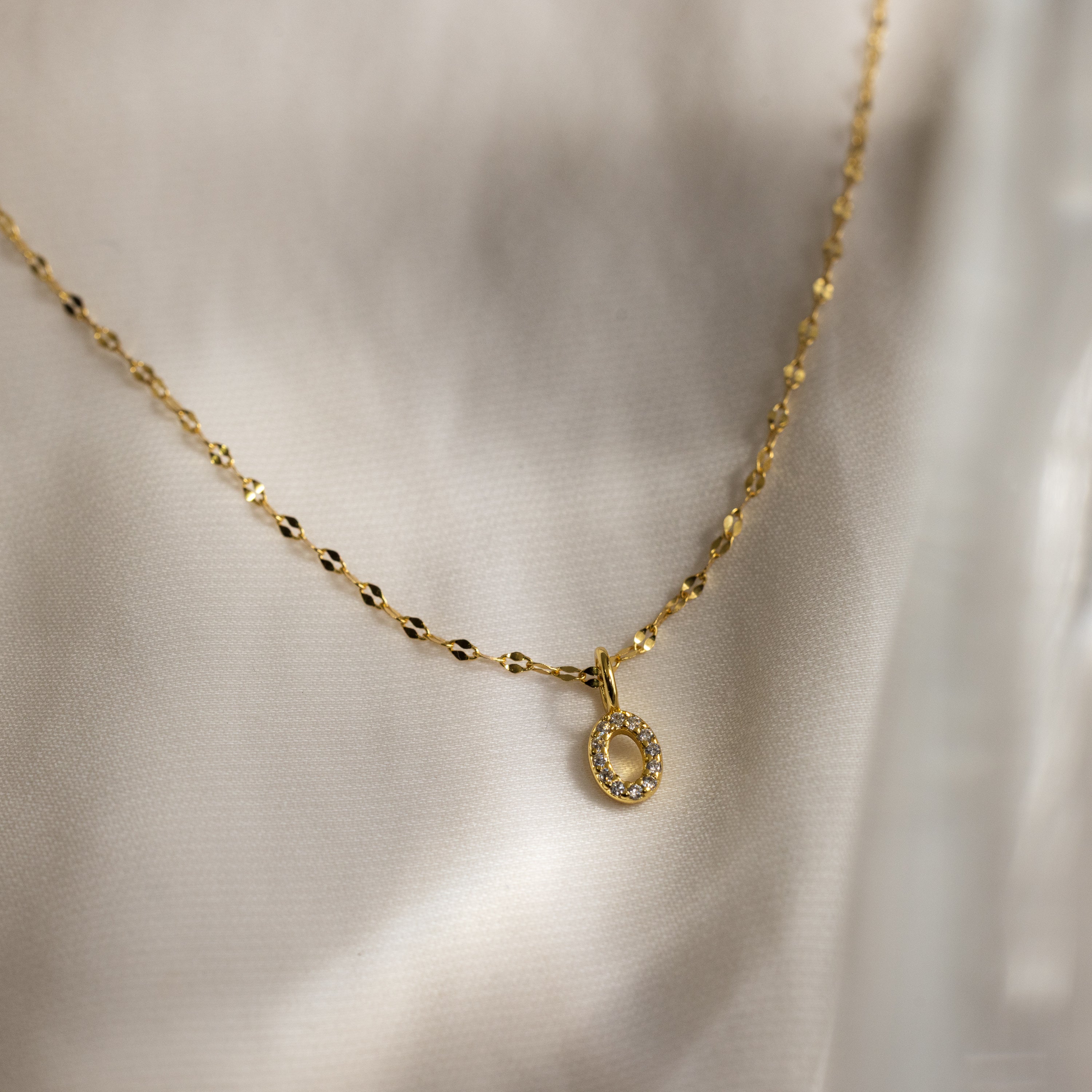Dainty Pave Letter Necklace in 18K Gold, featuring a small circular pendant with CZ diamonds, shown on a cream fabric background.