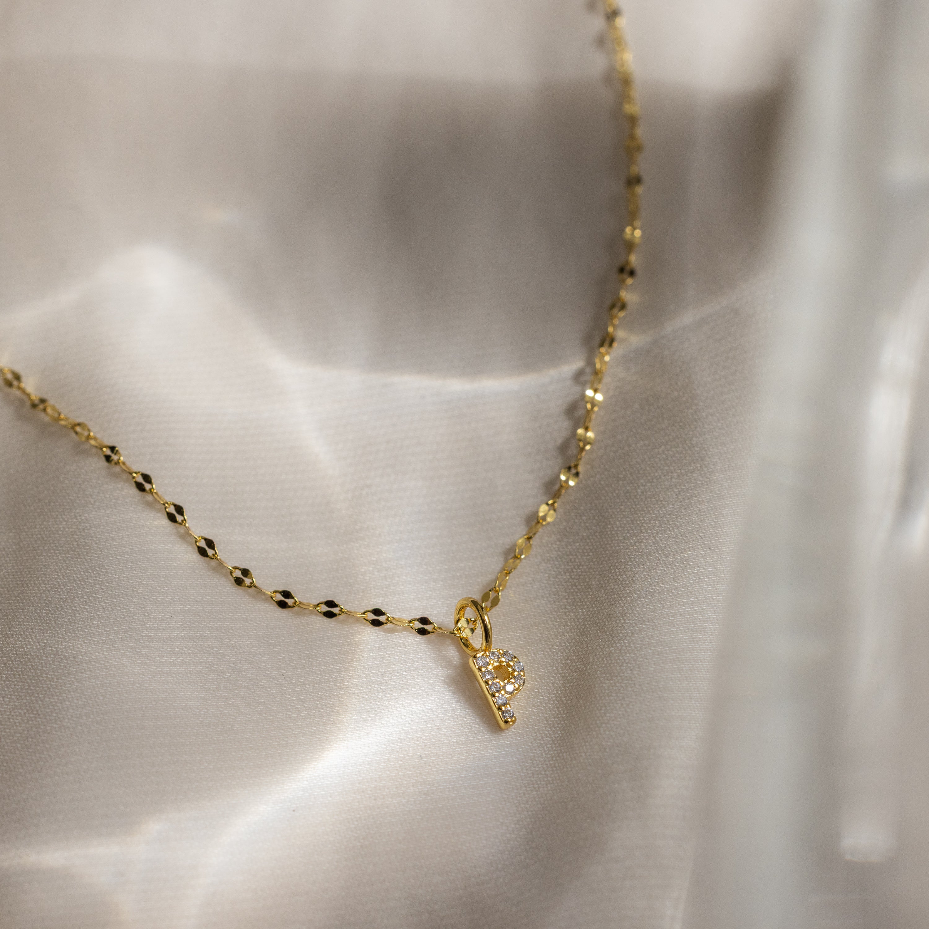 The Dainty Pave Letter Necklace in 18K Gold shines on silky, light-colored fabric, featuring a delicate gold chain and a sparkling pave letter pendant.
