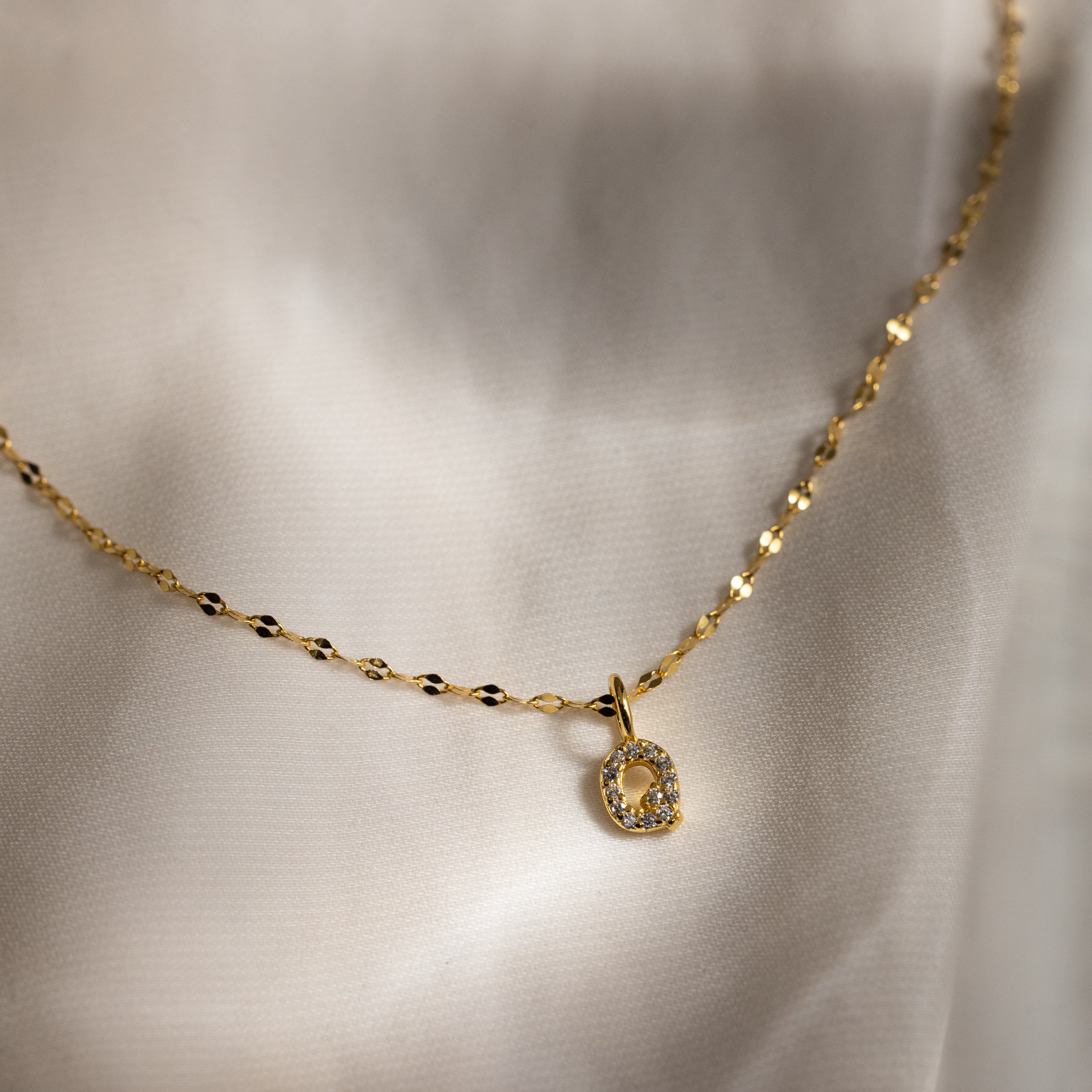 The Dainty Pave Letter Necklace in 18K Gold features a small round letter pendant encrusted with sparkling CZ diamonds, resting elegantly on white satin fabric.