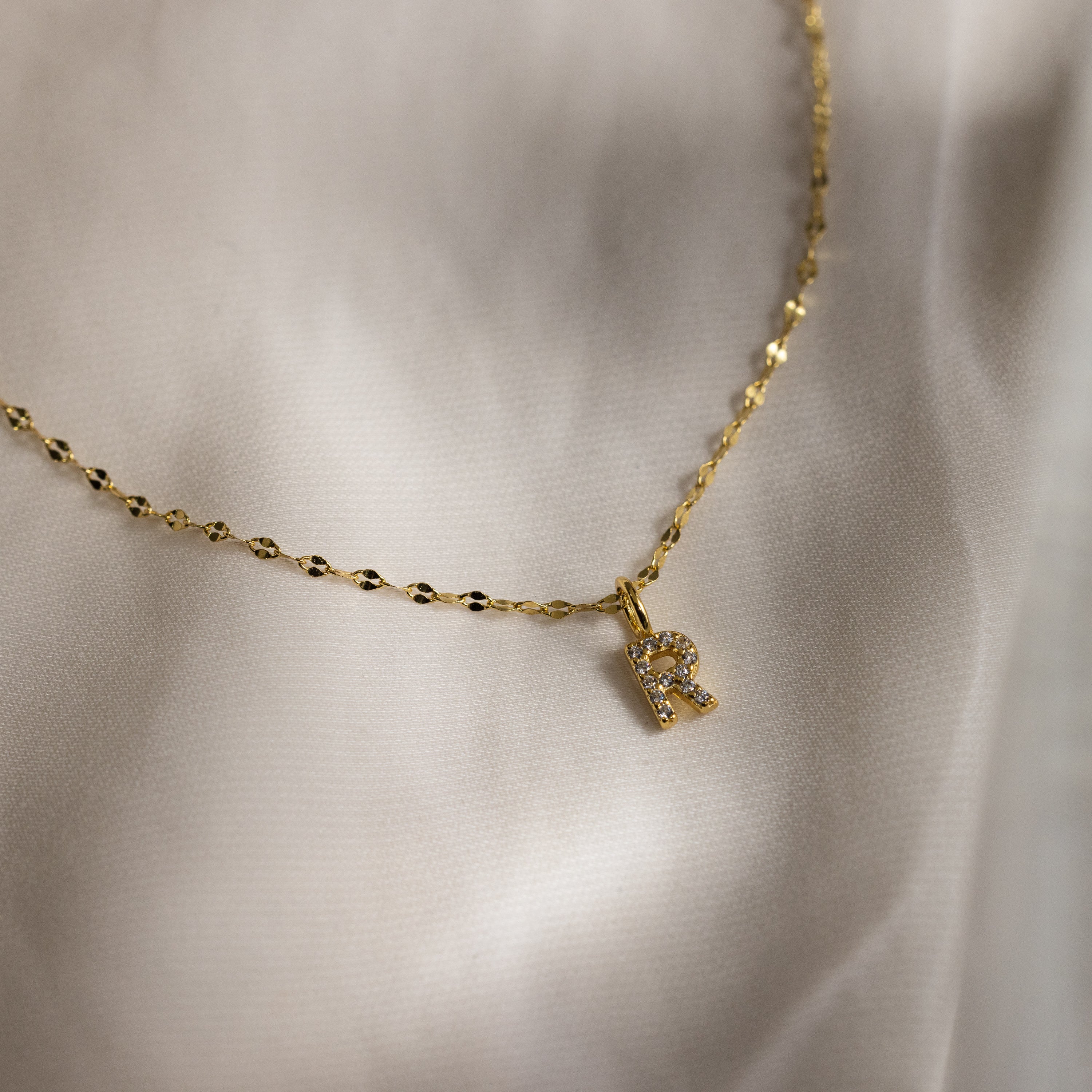 The Dainty Pave Letter Necklace in 18K Gold features a gold mirror chain and a pavé letter R pendant with sparkling CZ diamonds, showcased on a soft, light-colored fabric background.