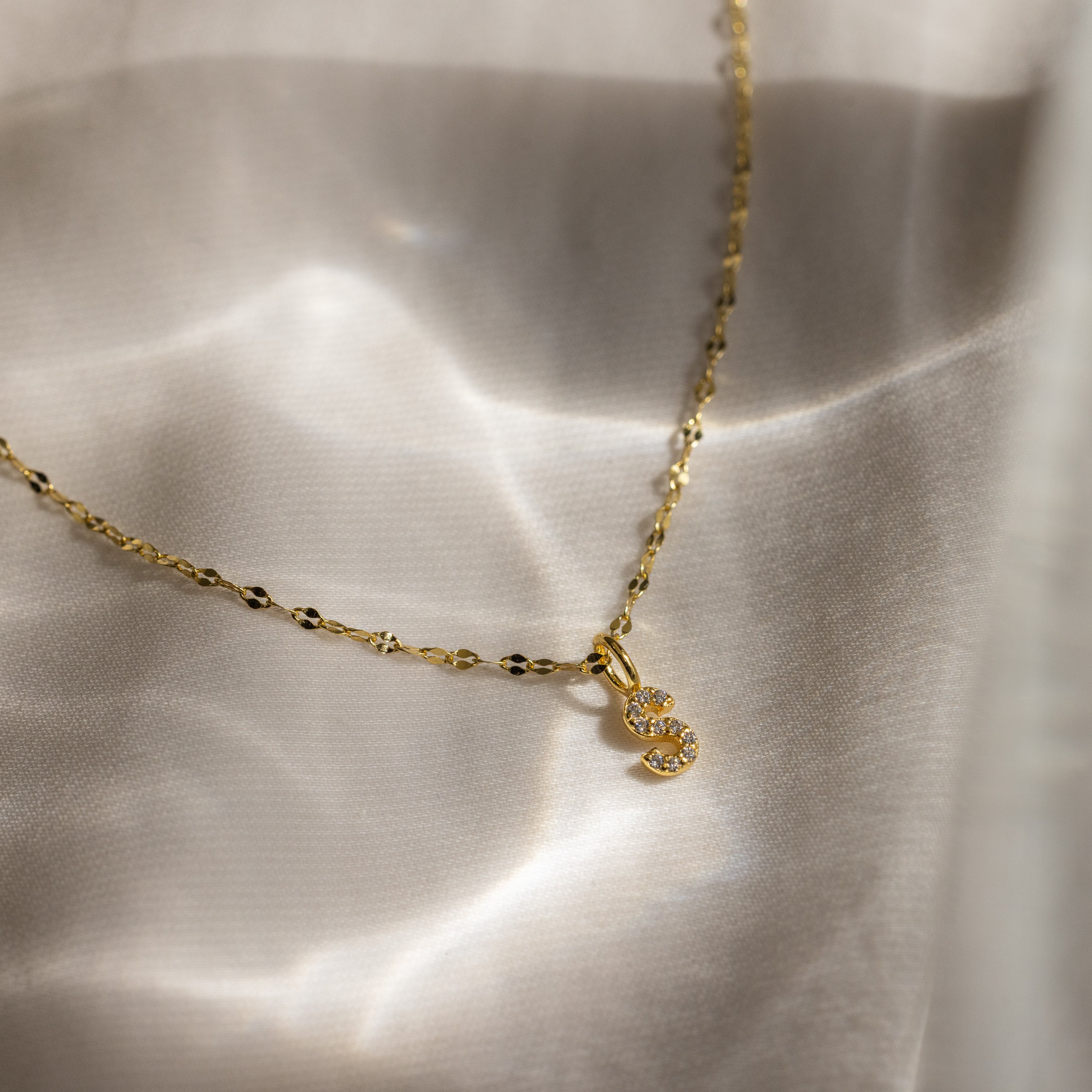 The Dainty Pave Letter Necklace in 18K Gold features a sparkling “S” pendant with CZ stones, beautifully displayed on soft, light-colored fabric under gentle sunlight.