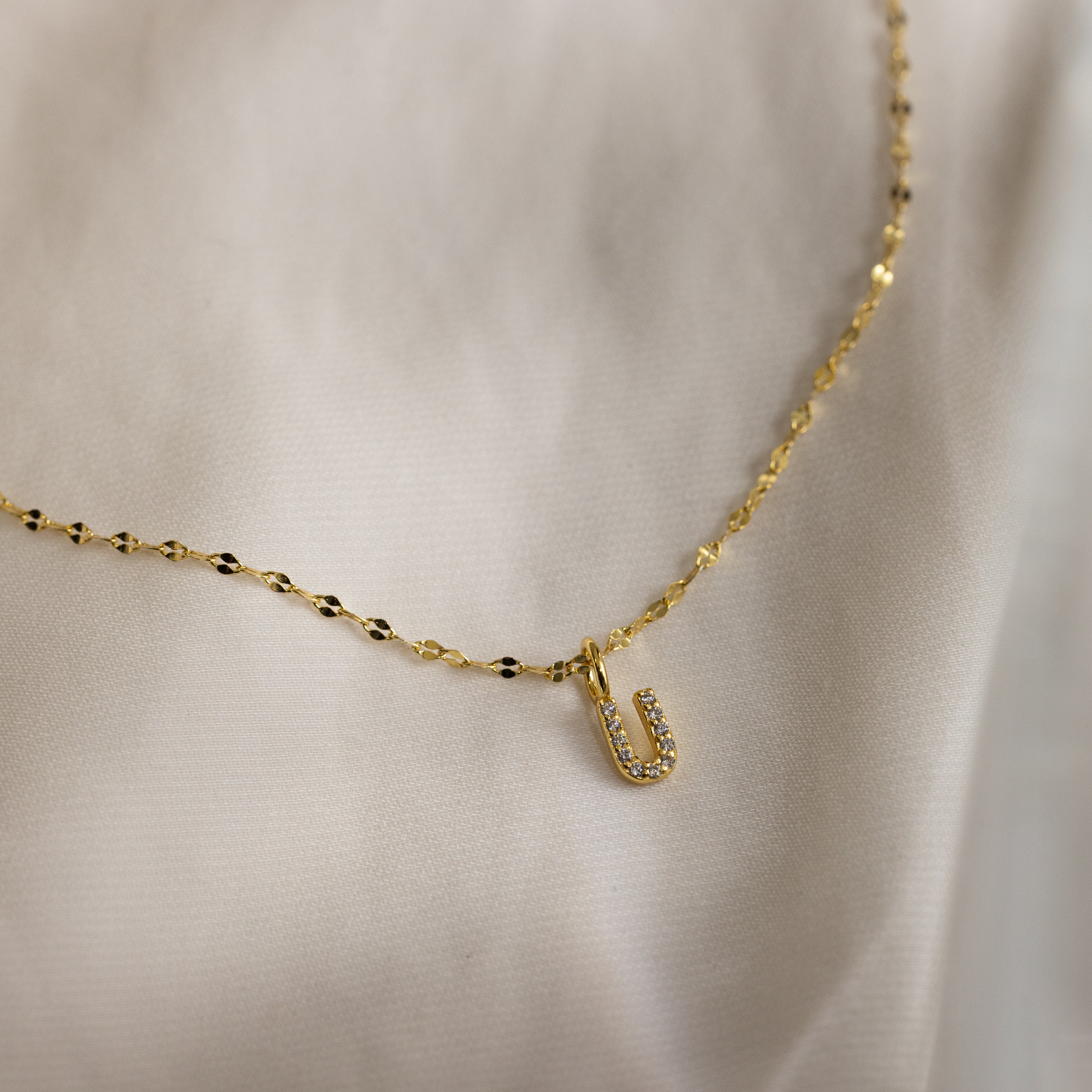 The Dainty Pave Letter Necklace in 18K Gold features a gold mirror chain with a small pavé letter U pendant adorned with CZ diamonds, displayed on soft, light-colored fabric.