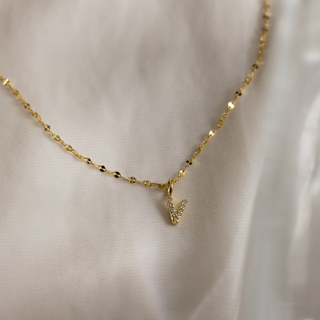 The Dainty Pave Letter Necklace in 18K Gold showcases a pavé letter V pendant with sparkling CZ diamonds on a gold chain, elegantly displayed against soft white fabric.