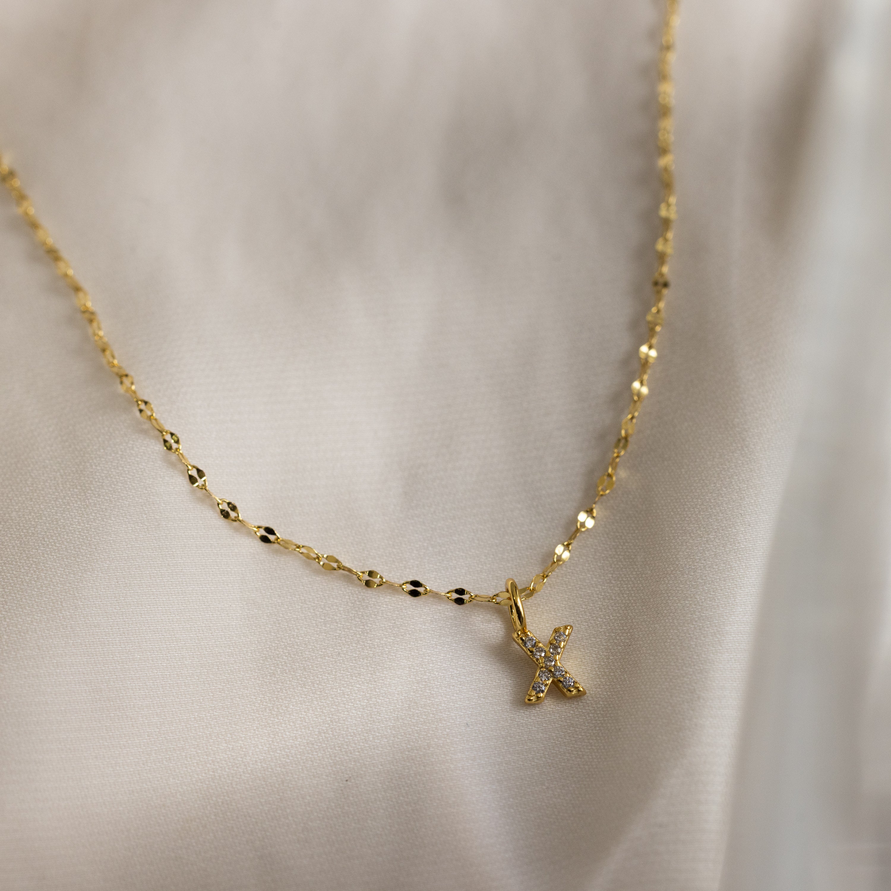 Dainty Pave Letter Necklace in 18K Gold featuring a small, CZ diamond-studded letter pendant on a delicate mirror chain, displayed on a light, silky fabric background.