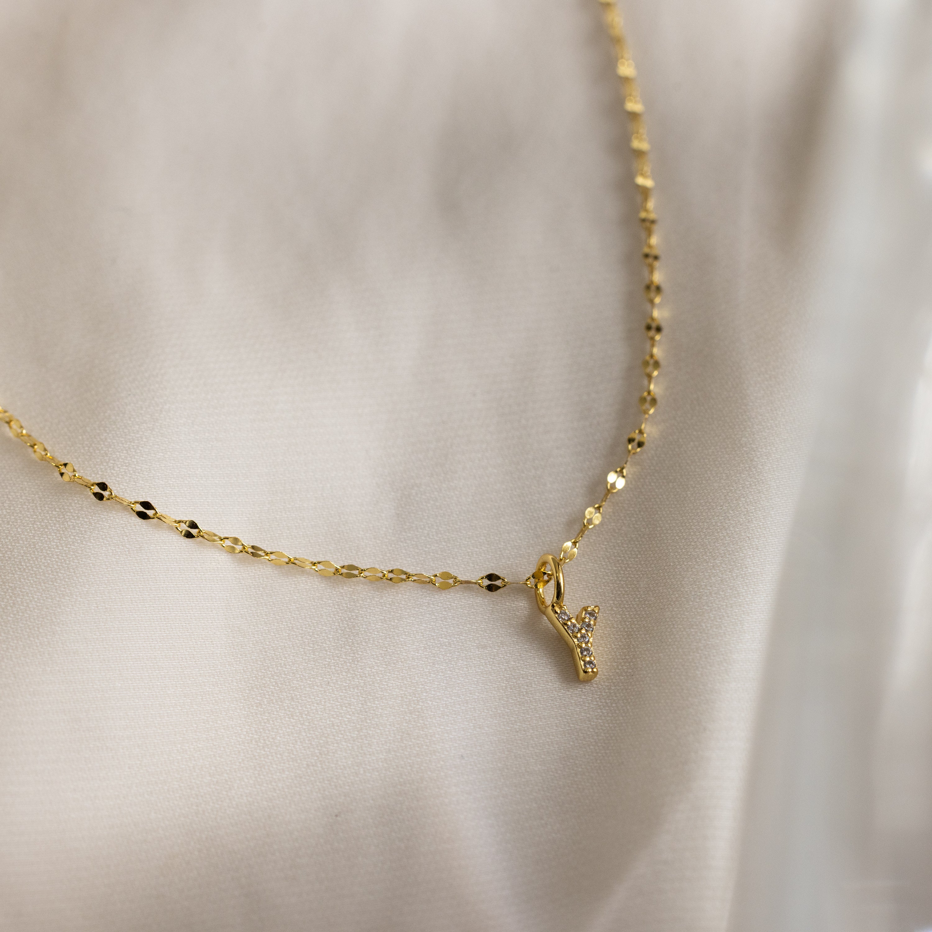 The Dainty Pave Letter Necklace in 18K Gold features a delicate mirror chain and a small, textured letter Y pendant adorned with sparkling CZ diamonds, showcased on soft, light-colored fabric.