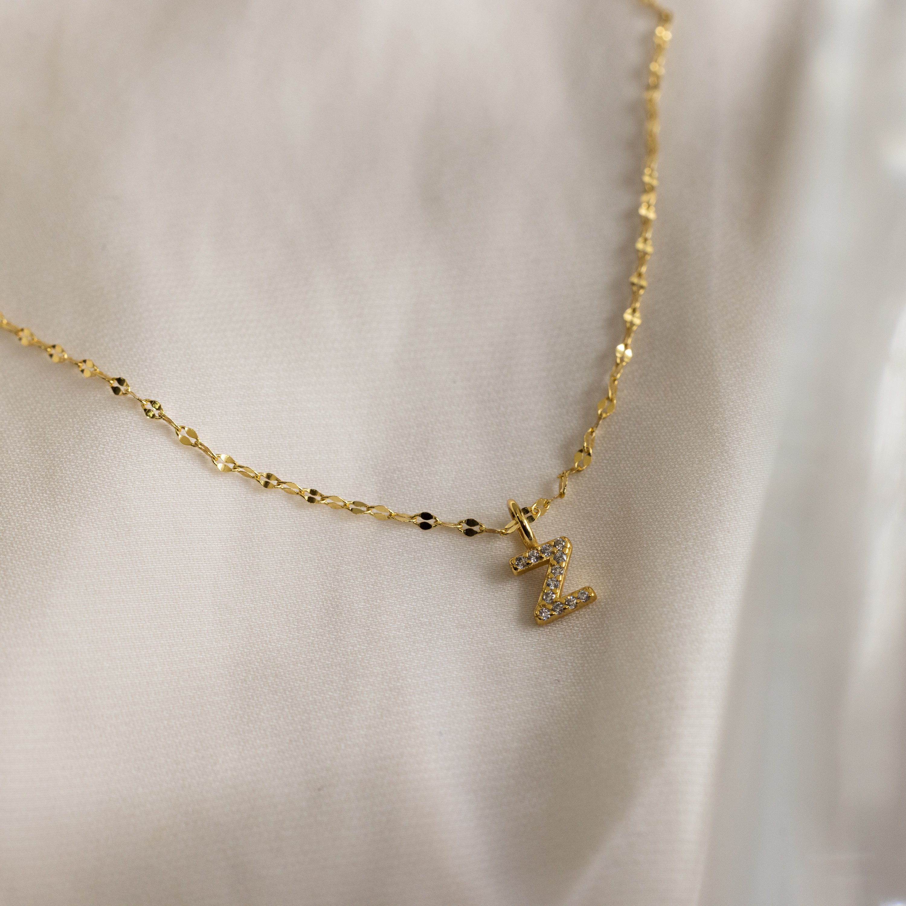 The Dainty Pave Letter Necklace in 18K Gold features a textured mirror chain and a small pavé letter Z pendant with sparkling CZ diamonds, elegantly displayed on soft fabric.