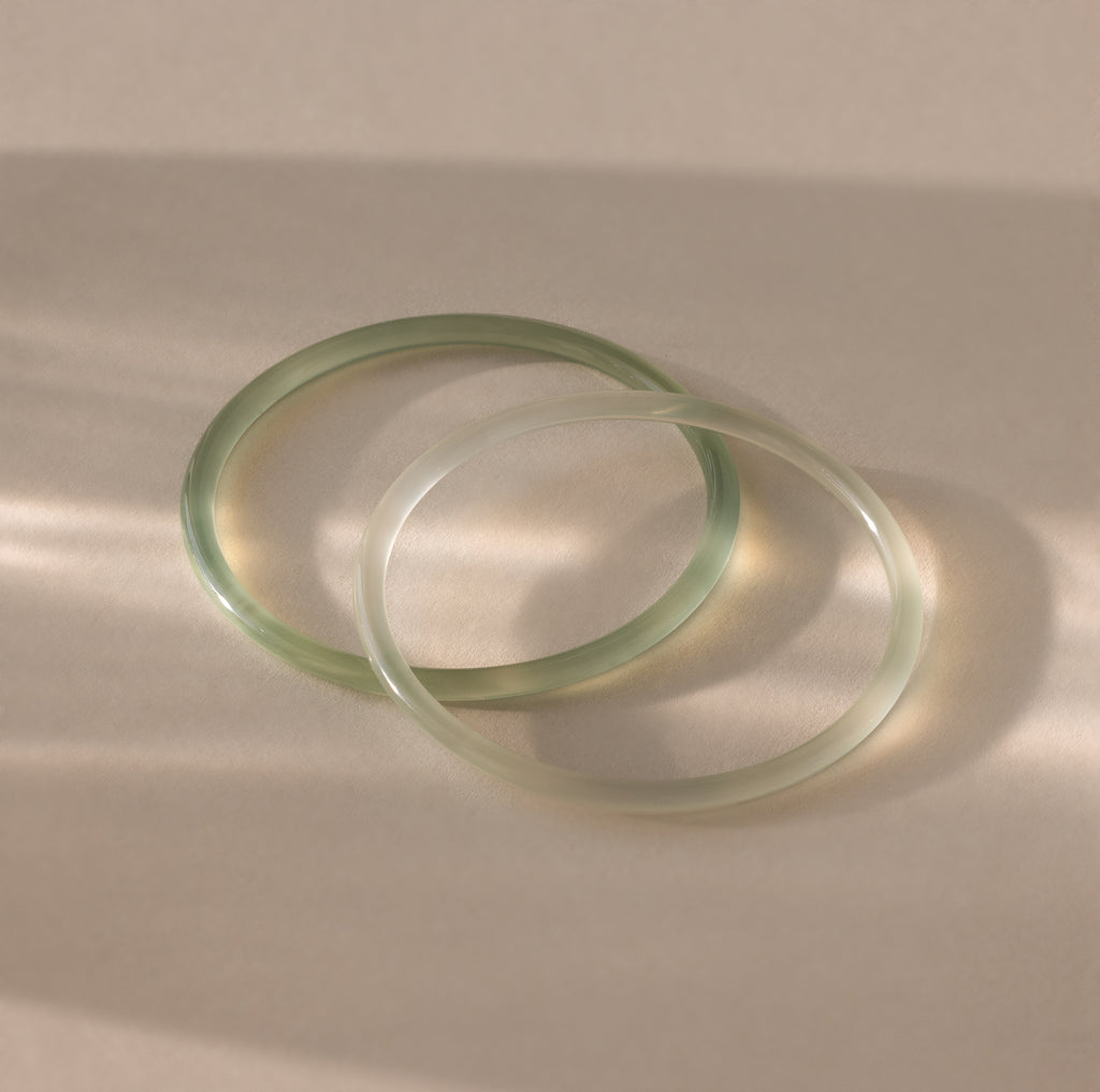 Close-up of two thin jade bangles in soft translucent green tones, displayed on a neutral surface.