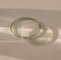Close-up of two thin jade bangles in soft translucent green tones, displayed on a neutral surface.