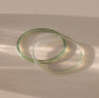 Close-up of two thin jade bangles in soft translucent green tones, displayed on a neutral surface.