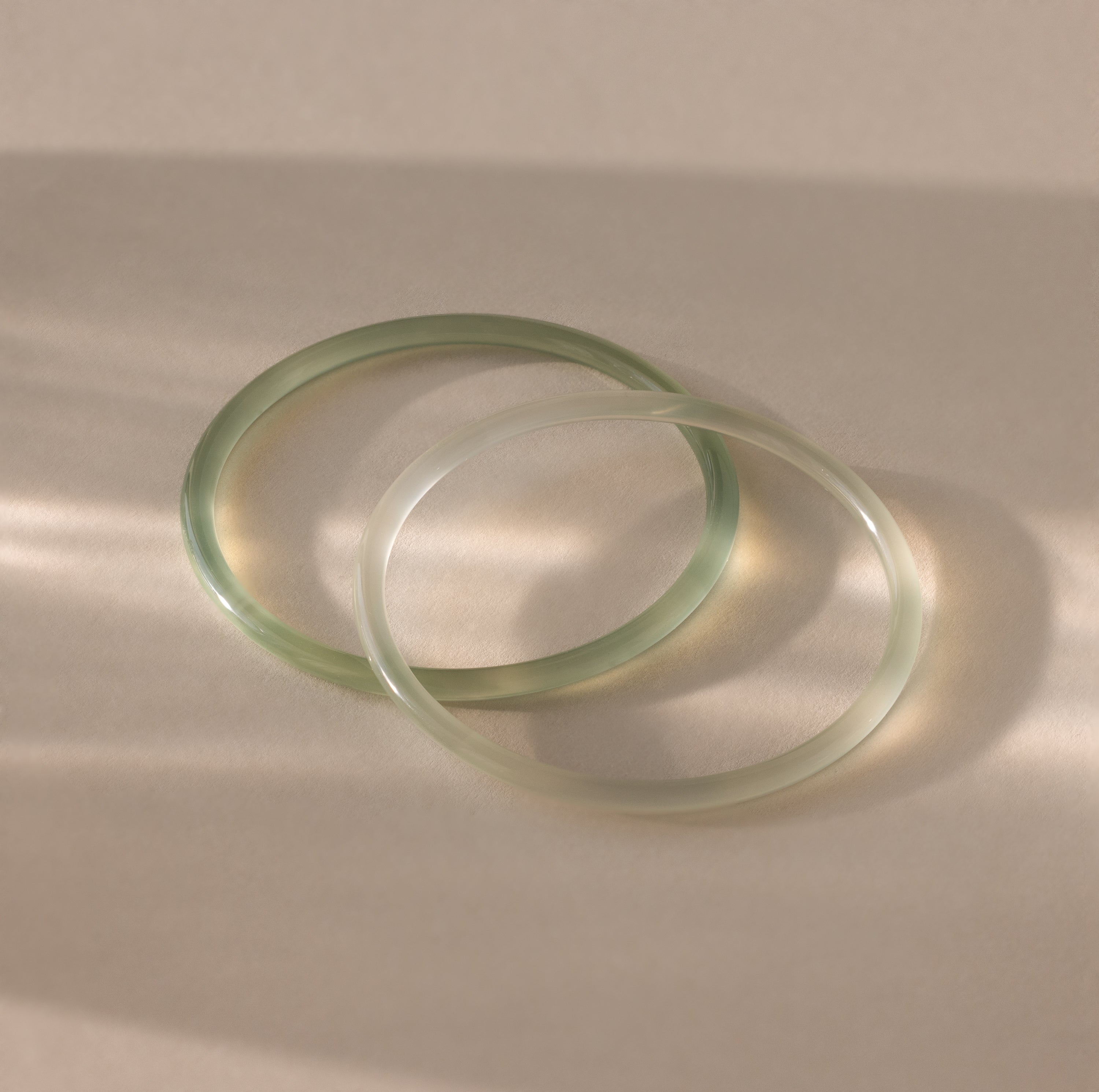 Close-up of two thin jade bangles in soft translucent green tones, displayed on a neutral surface.