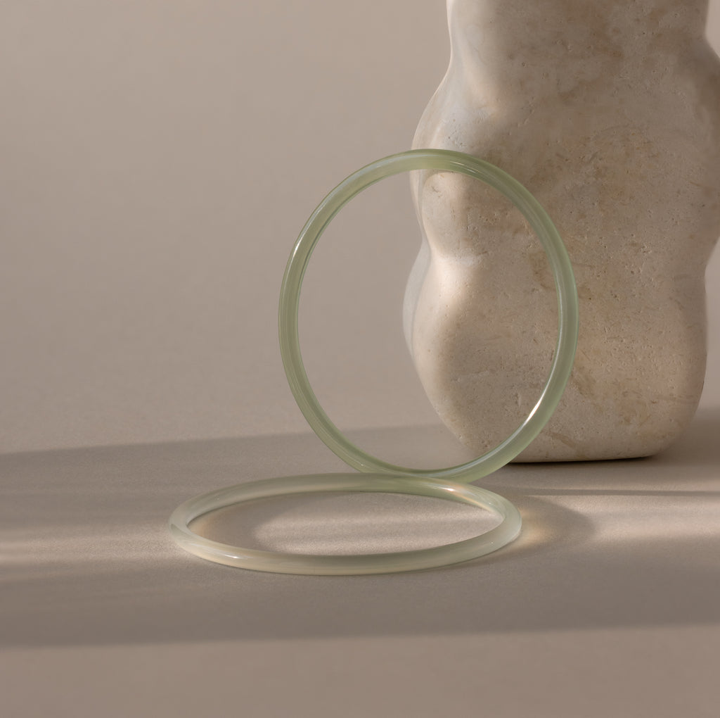 Set of two thin jade bangles in soft translucent green tones, with the darker of the two resting up-right against a stone prop while the lighter bracelet lays flat.
