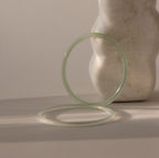 Set of two thin jade bangles in soft translucent green tones, with the darker of the two resting up-right against a stone prop while the lighter bracelet lays flat.